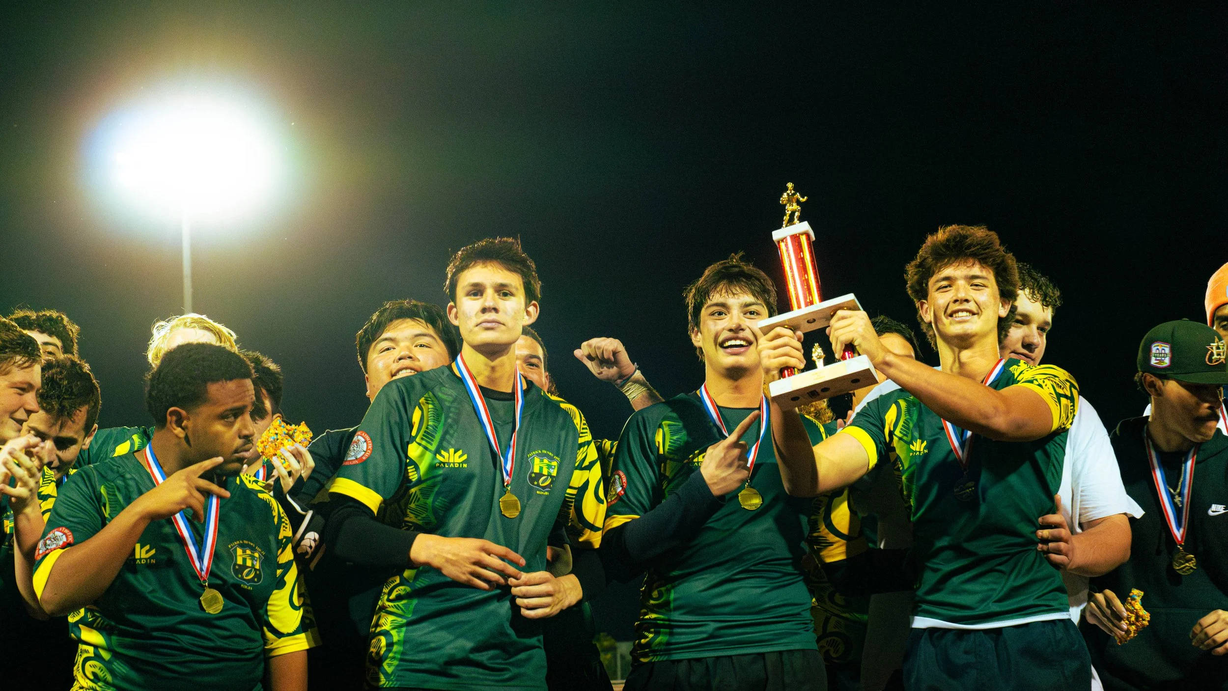 A group of young male athletes in green and yellow uniforms celebrating a win on a sports field under stadium lights, with some wearing medals and one holding a trophy.