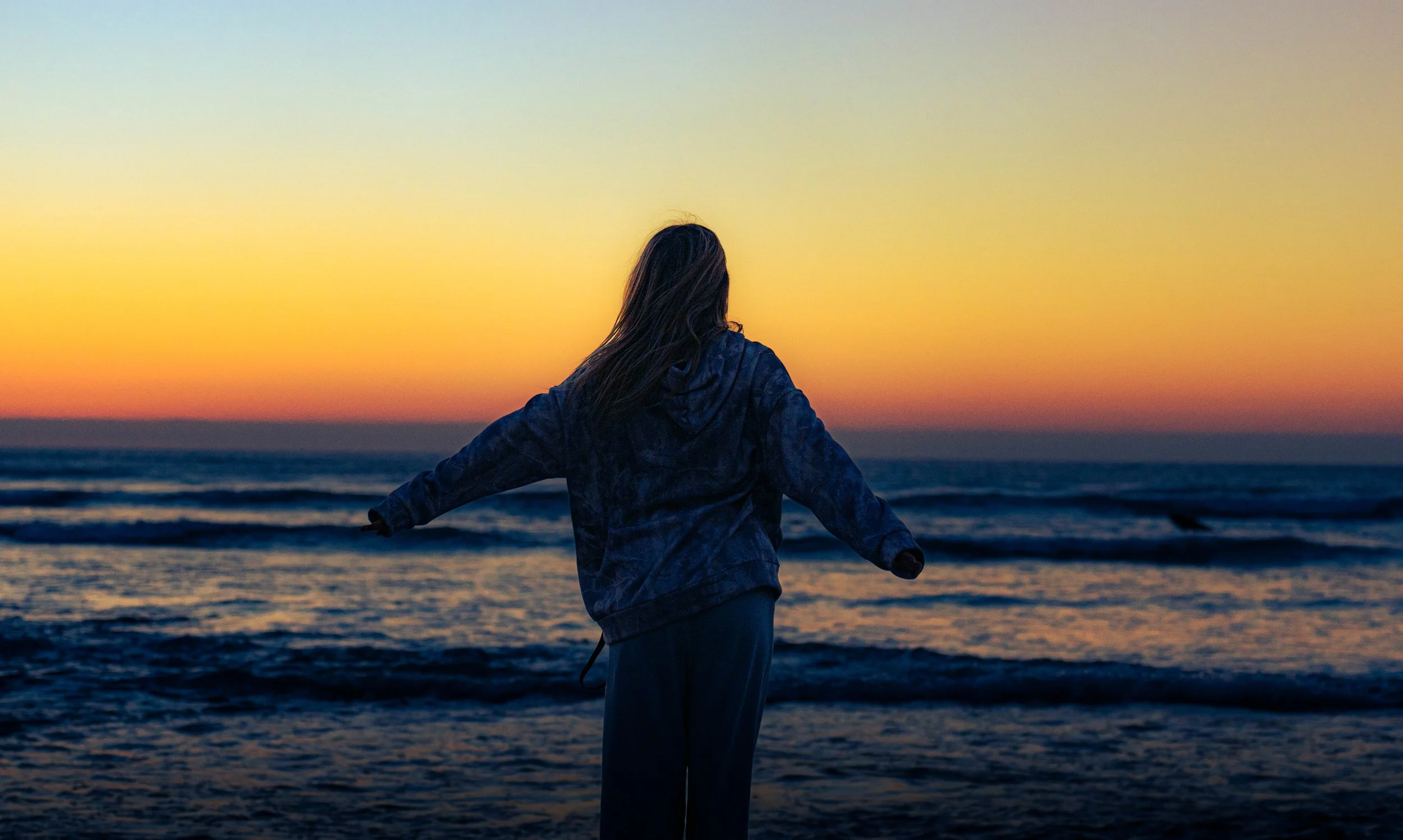 Person with long hair wearing a jacket standing on a beach at sunset, facing the ocean with arms outstretched.
