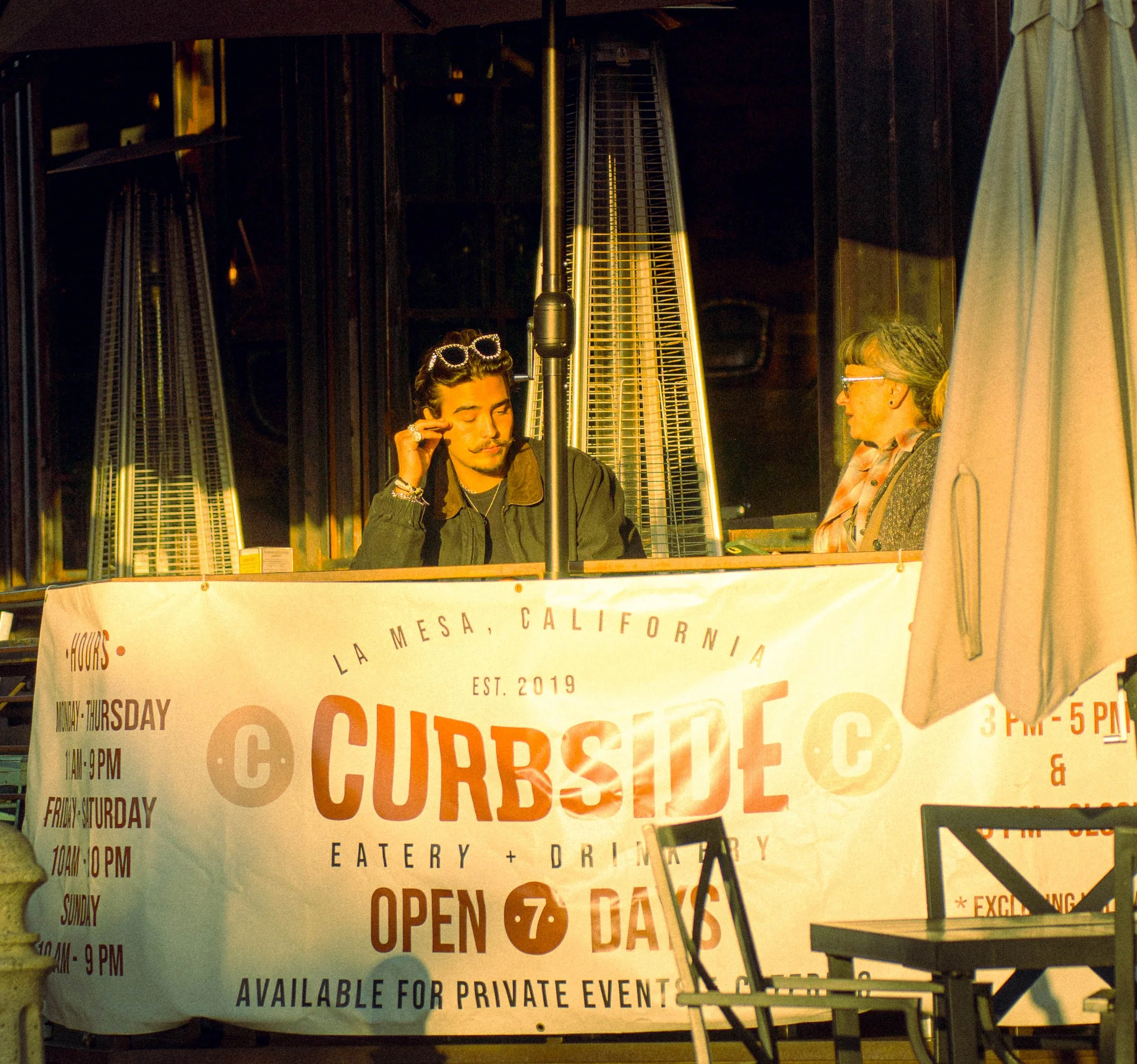 A group of people sitting outdoors at a table on a patio, with a large heater and a banner advertising a restaurant or food event in La Mesa, California, during evening hours.