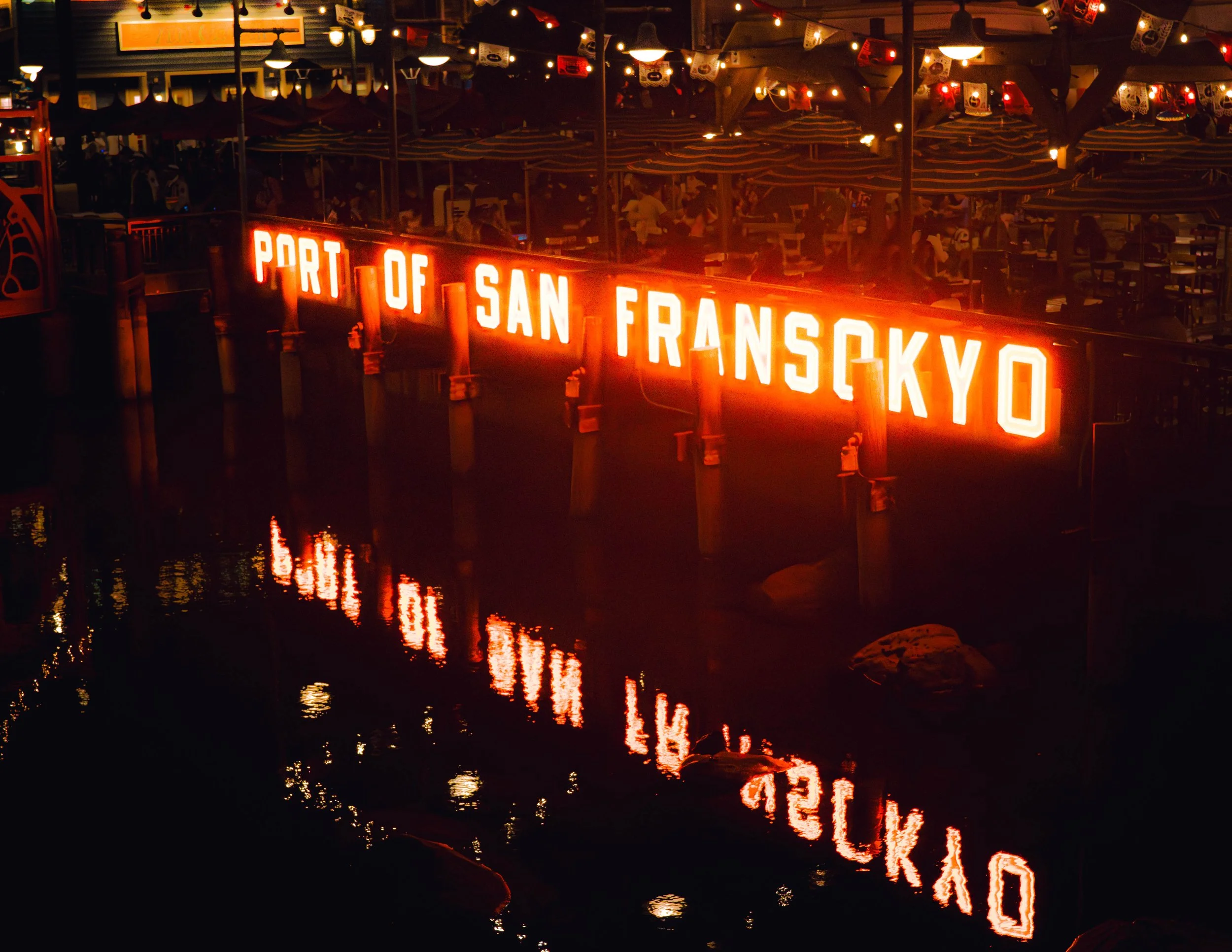 Nighttime scene at a port with a large, illuminated sign reading 'PORT OF SAN FRANCISCO' reflected in the water, with tables, umbrellas, and string lights visible in the background.
