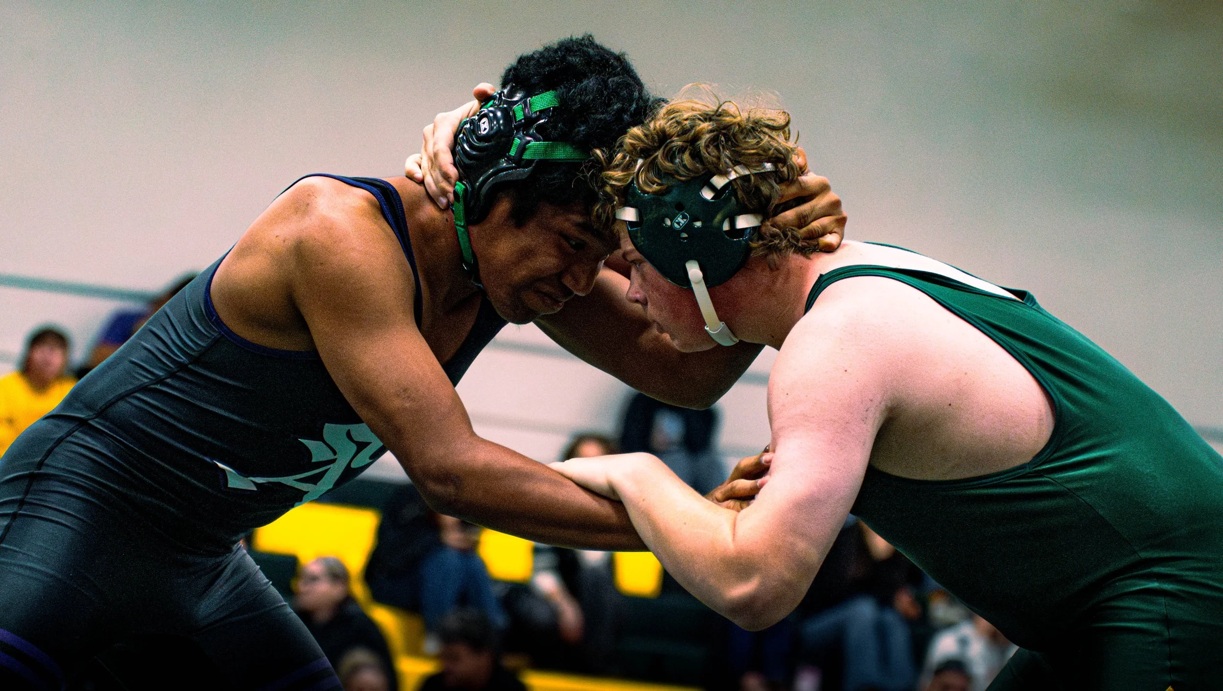 Two wrestlers in singlets and protective headgear engage in a match, gripping each other's arms and bearing down on each other.