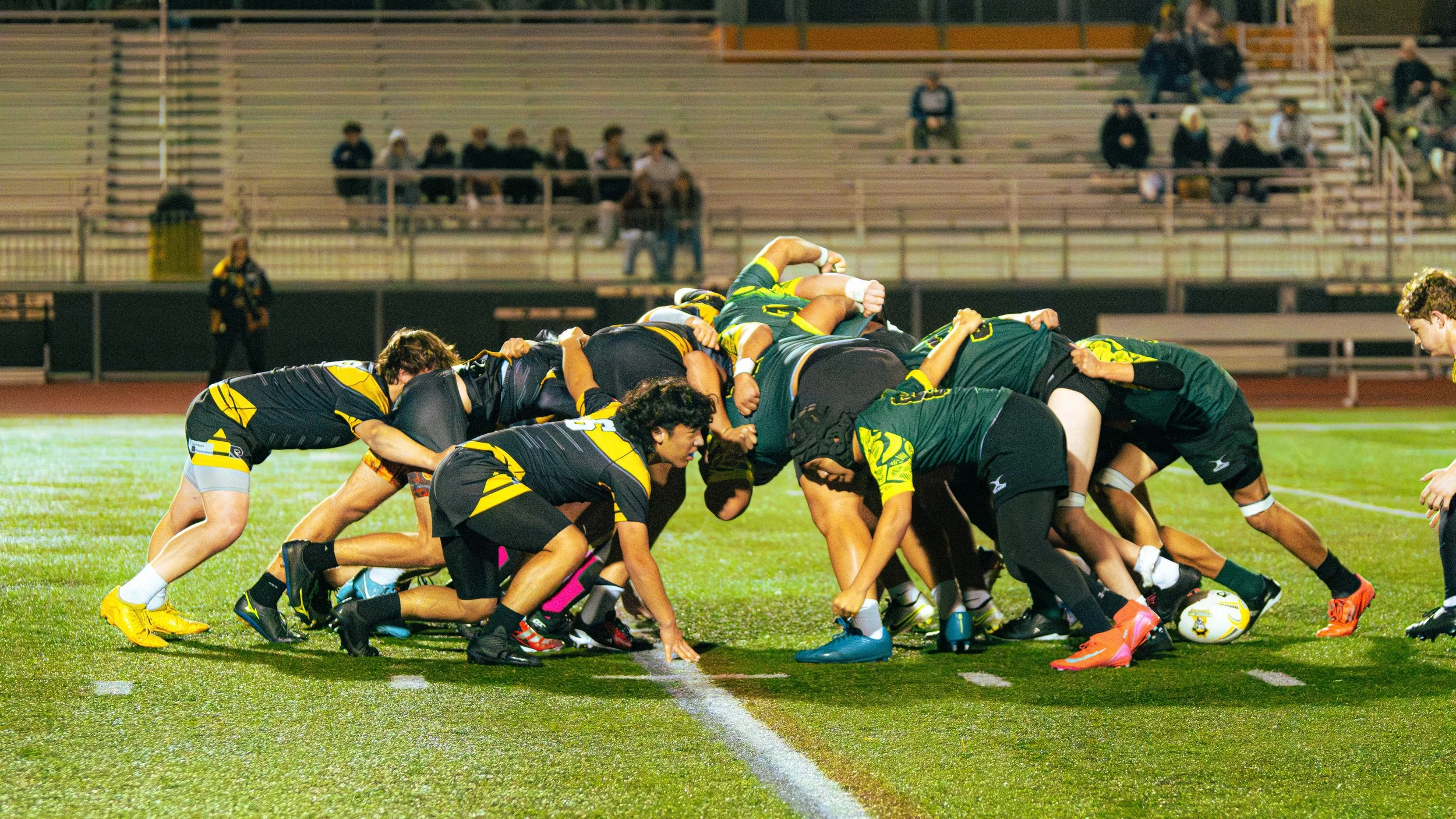 A rugby team in black and yellow uniforms battles for possession of the ball during a nighttime game on a wet field, with spectators seated in the background.