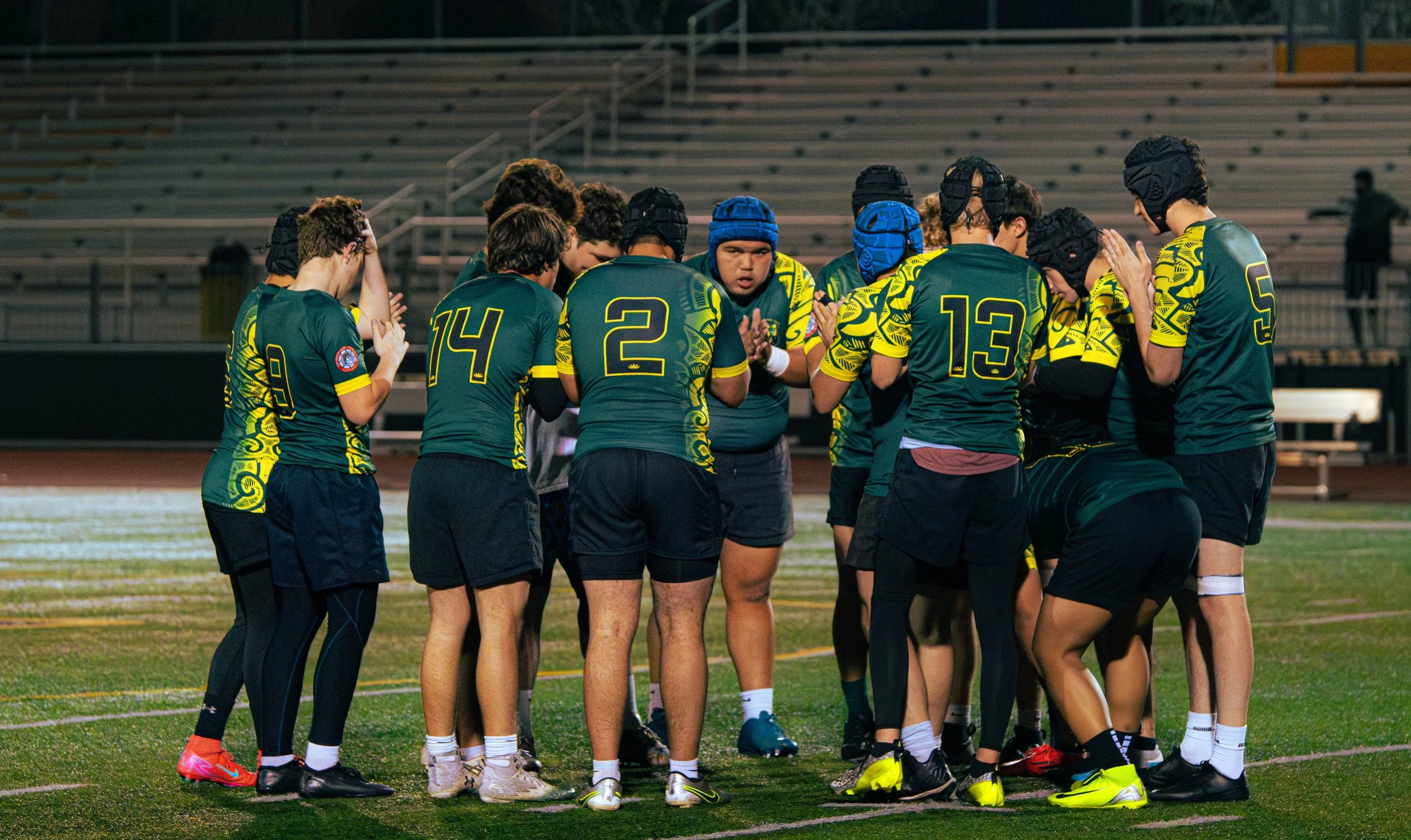 A group of male rugby players wearing green and yellow jerseys, black shorts, and helmets, gathered in a huddle on a rugby field at night.