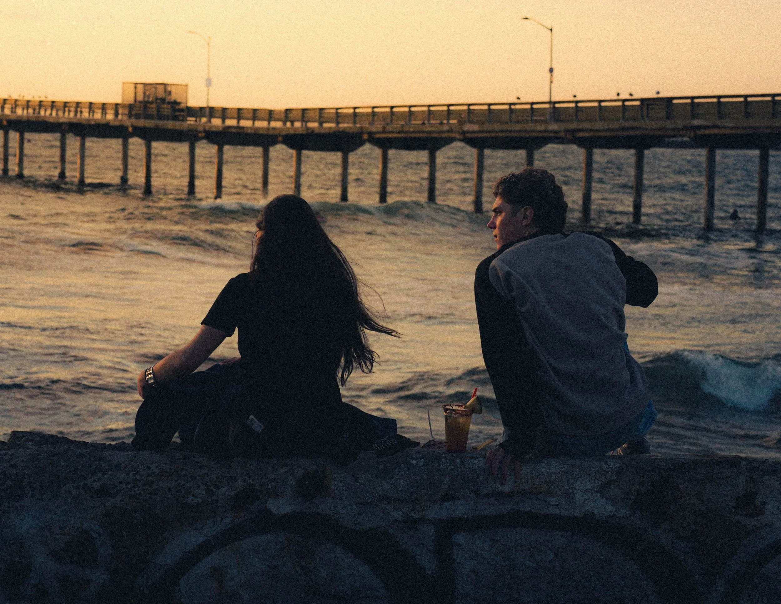A young man and woman sit by the ocean at sunset, facing the water near a pier extending into the sea. The woman has long dark hair, and the man has short, curly hair. They are casually dressed, and there is a drink with a straw and a slice of fruit placed on a rock in front of them.