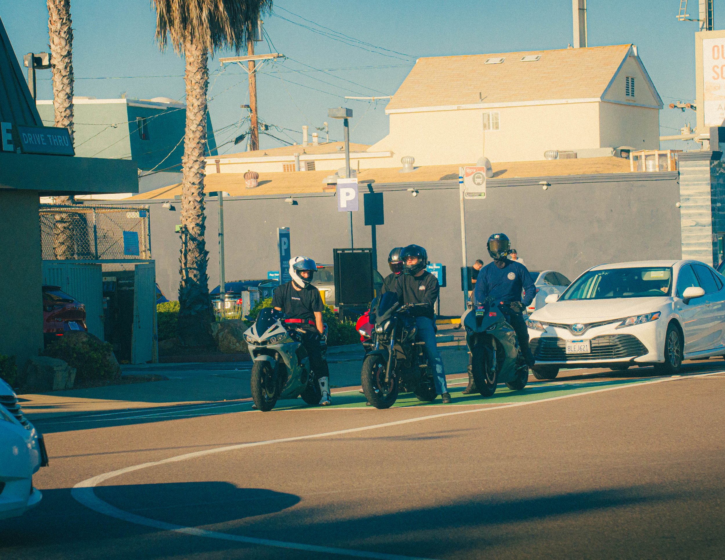 People wearing helmets sitting on motorcycles in a parking lot near cars, with a shaded building and palm trees in the background during daytime.