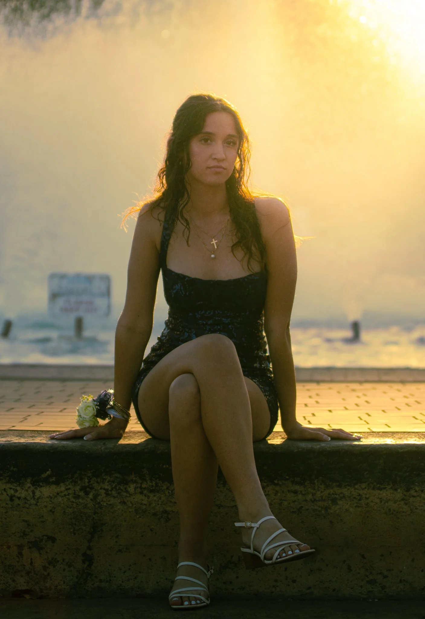 Woman sitting on a ledge with her legs crossed, wearing a black dress, white sandals, jewelry, and holding a bouquet of white roses, with sunset in the background.
