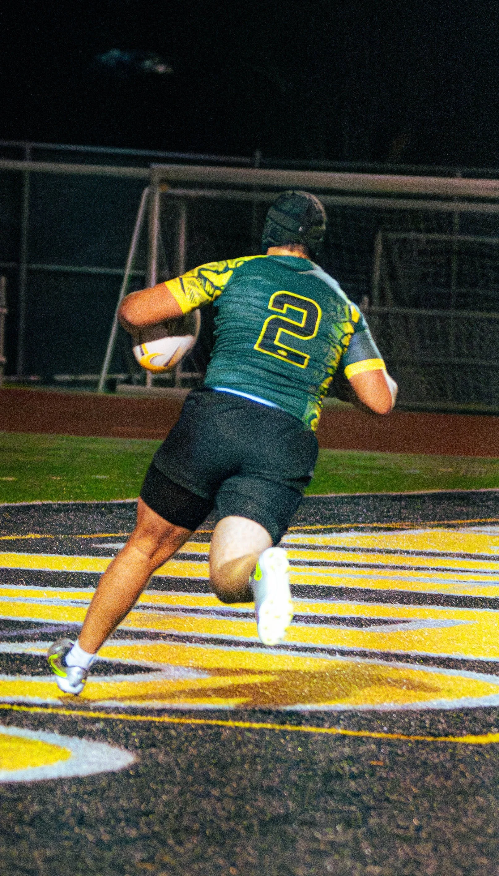 A football player wearing a dark uniform with the number 2 on his back running on a football field at night, holding a football in his right arm.