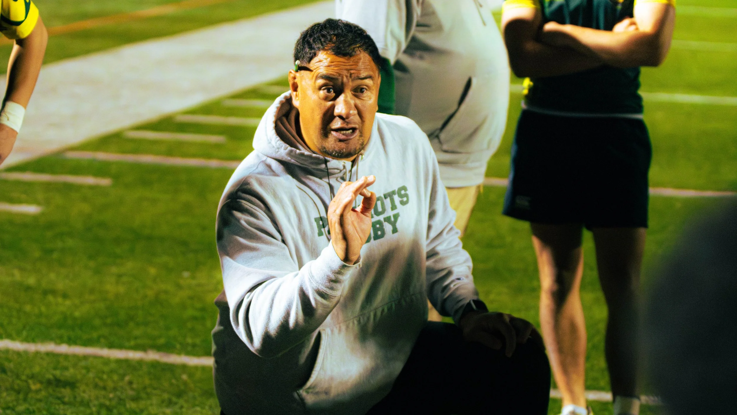 A coach or trainer speaking during a sports practice or game on a football field at night, with other players and individuals standing nearby.