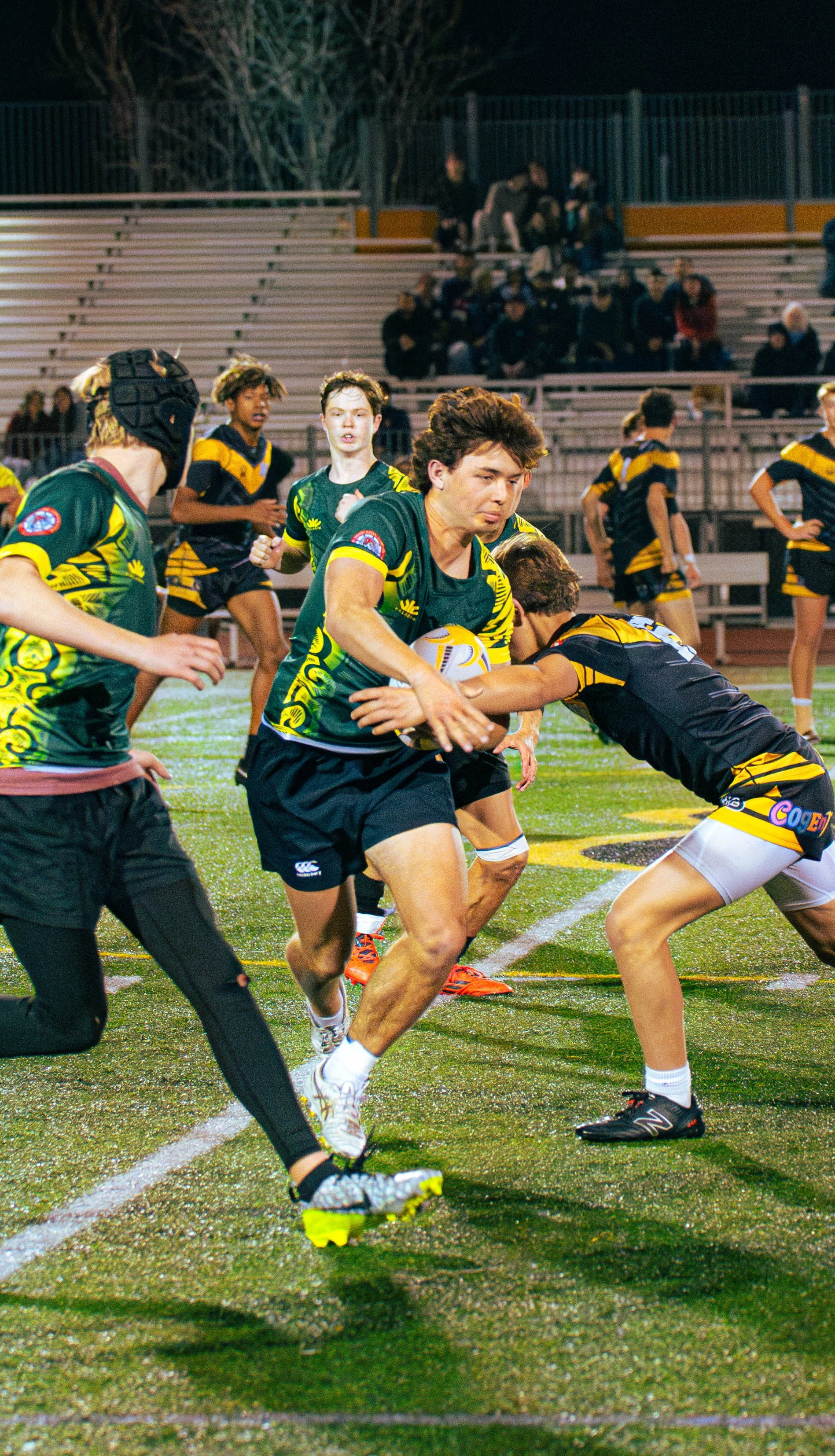 Nighttime rugby game with players in green and black uniforms, some wearing headgear, on a wet field with spectators watching from the bleachers.