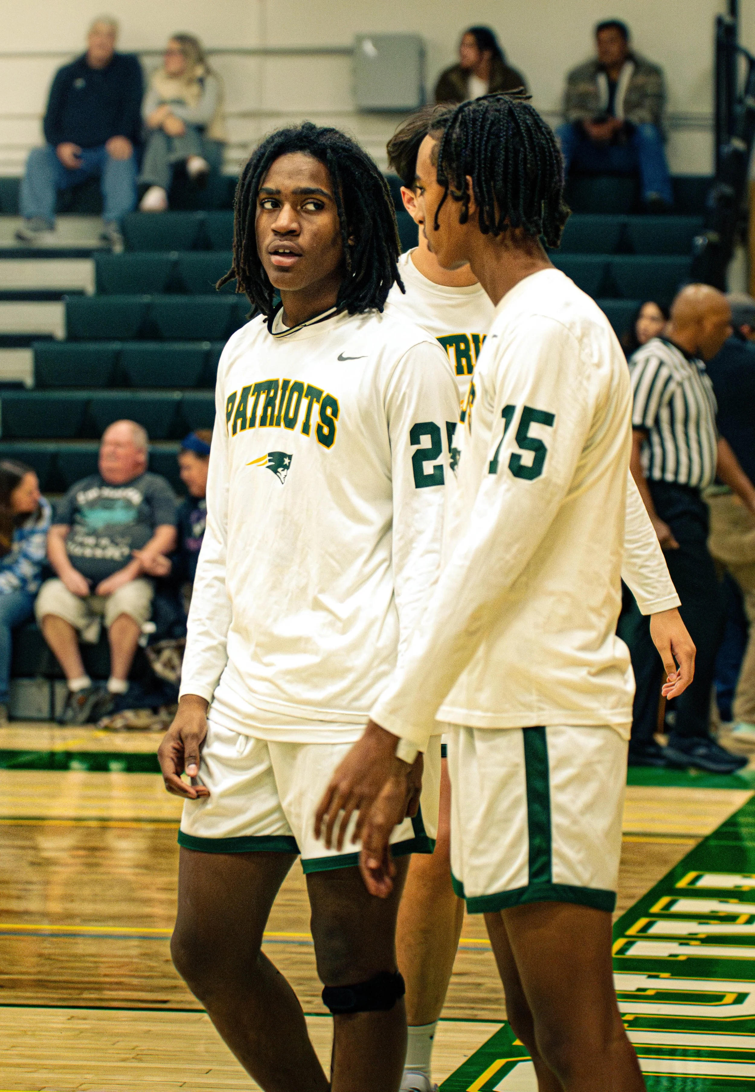 Two basketball players standing on the court during a game, wearing white jerseys with green accents and the word "Patriots." One player is looking at the camera with a surprised expression, and the other is facing away, appearing to be mid-conversat