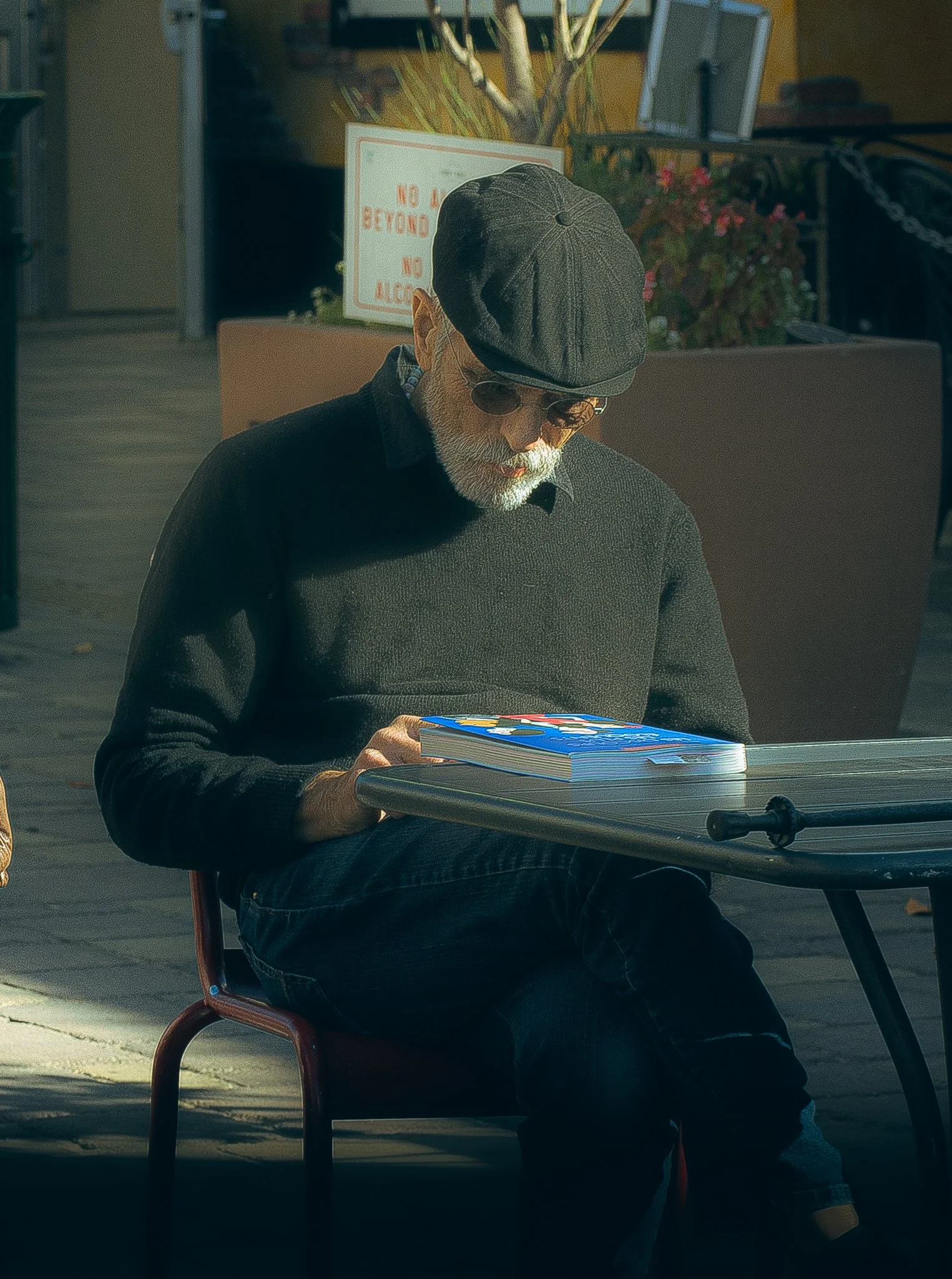 An elderly man with a white beard, wearing glasses, a black cap, and a black sweater, seated outdoors at a small table, reading a colorful comic book. Sunlight falls on him, and there are plants and a sign that reads 'No' in the background.