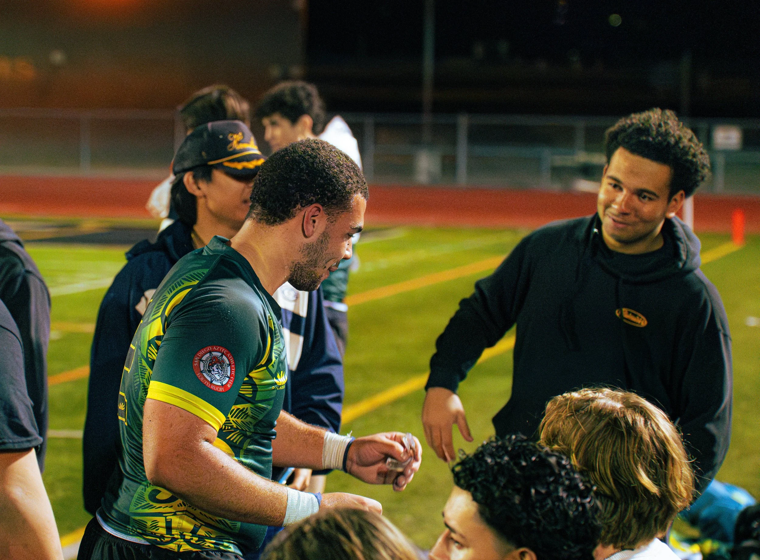A group of young people on a sports field at night, interacting and talking, with some wearing athletic clothing. One person in the foreground is wearing a green and yellow sports jersey, and others are engaged in conversation.