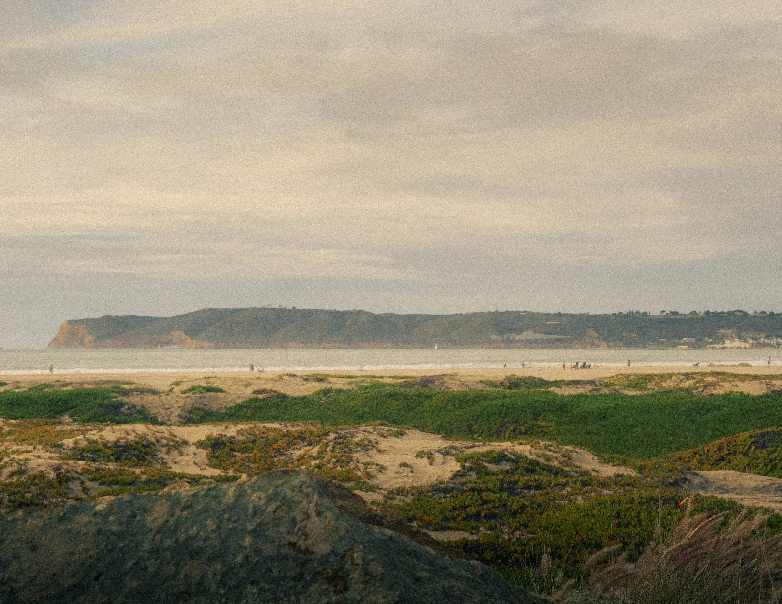 A view of a cloudy sky over a coastal scene with rocky cliffs, a sandy beach, and some patches of greenery in the foreground. There are a few people walking along the beach and a sailboat in the water.