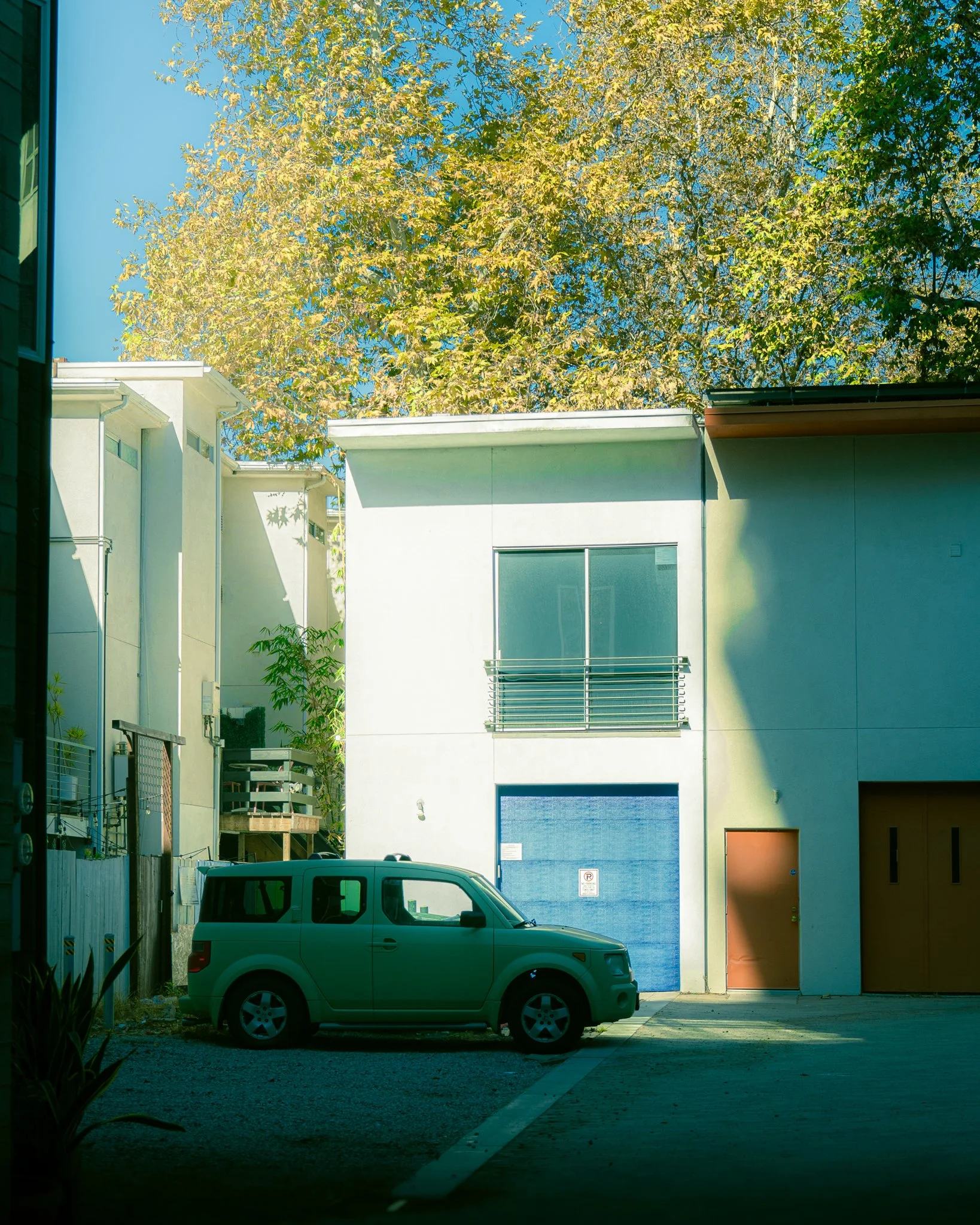 Modern multi-story building with light-colored exterior walls, large blue garage door, and various windows, with a silver SUV parked in front under a clear blue sky.