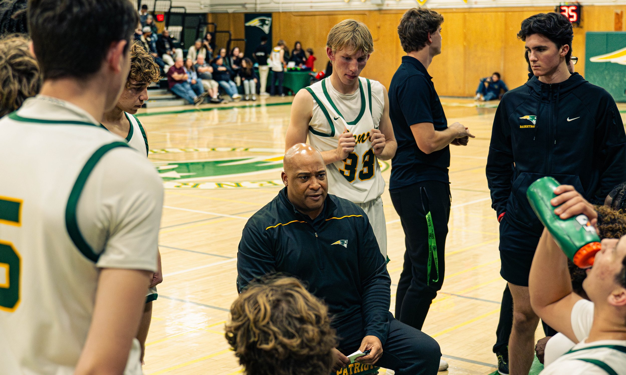 High school basketball team huddle during game, coach speaking, players listening, gymnasium with spectators in background.