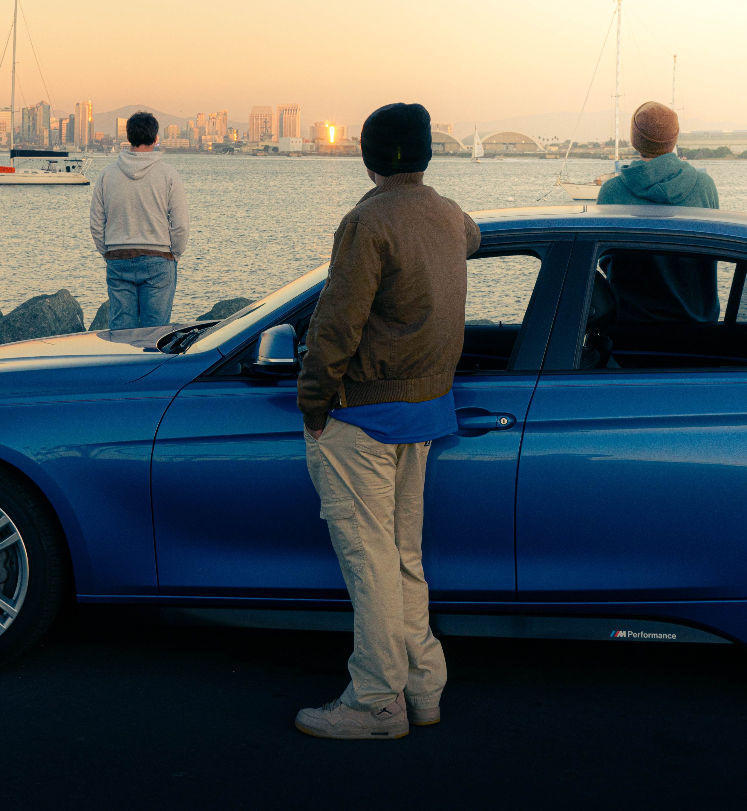 Three young men standing by a parked blue BMW car beside water, with a city skyline and sailboats in the background during sunset.