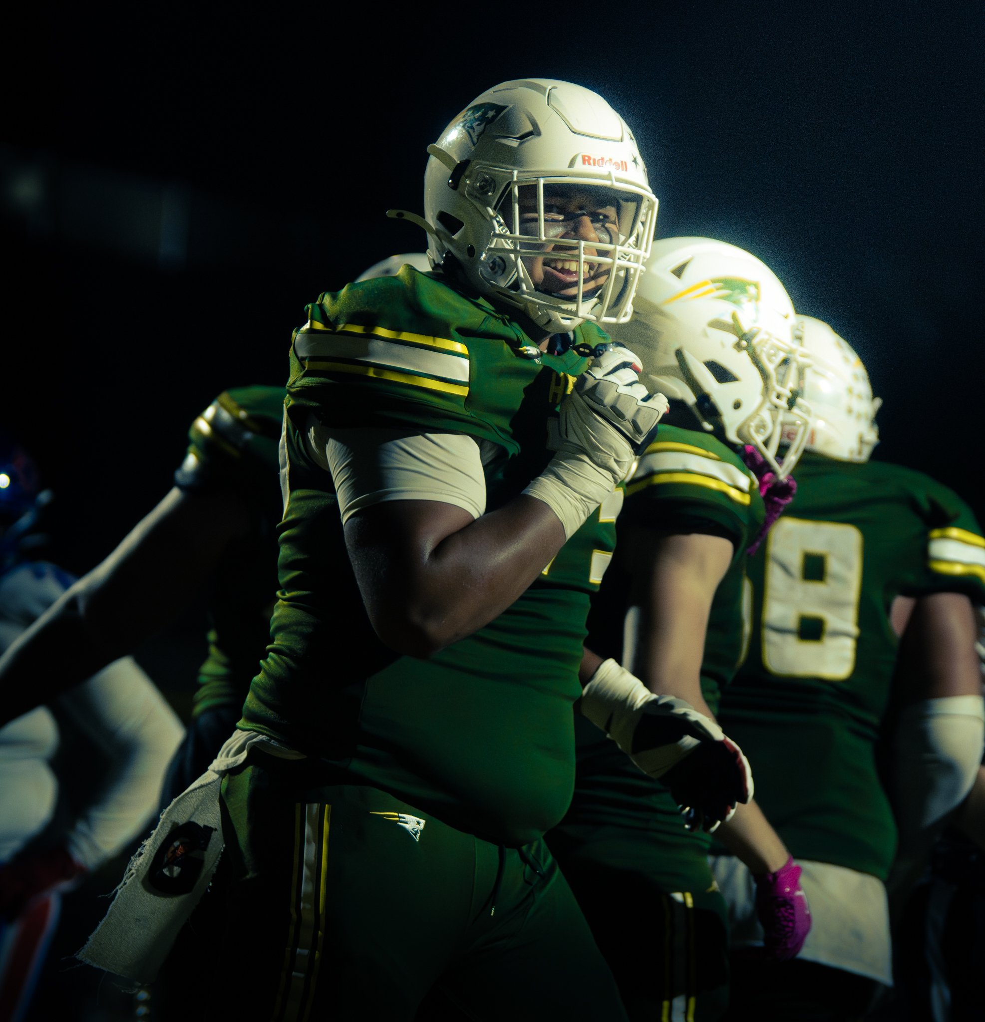 Football players in green and white uniforms, wearing helmets, standing in a huddle on the field during a night game.
