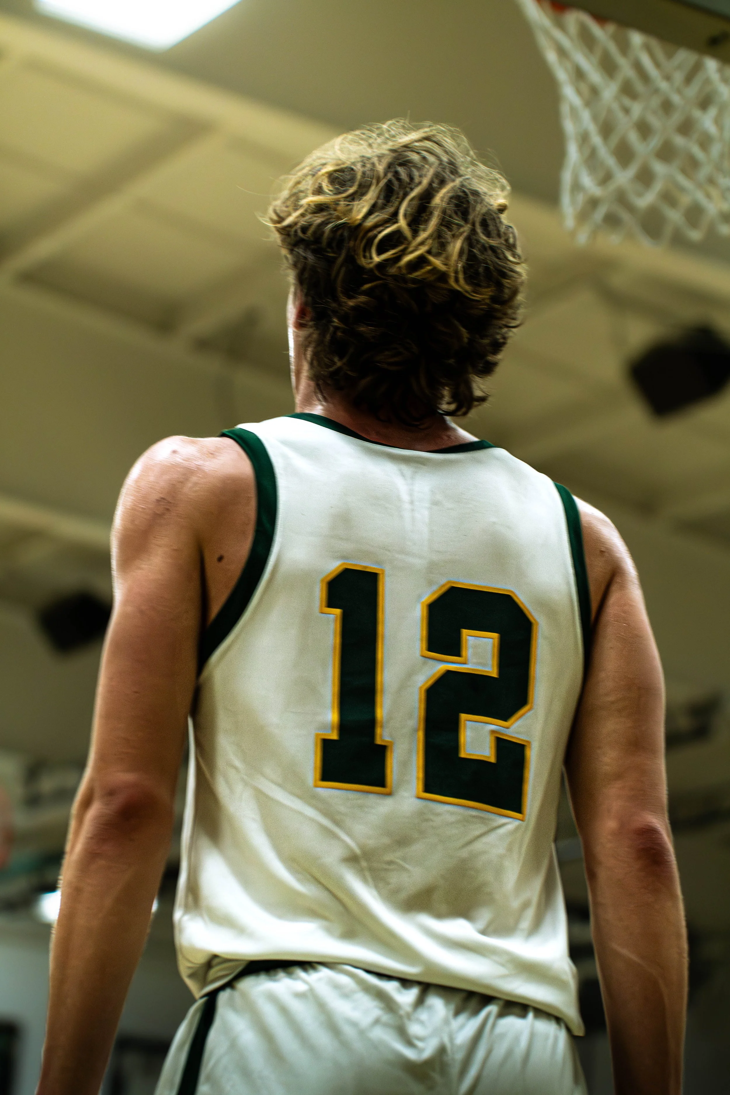 A female basketball player viewed from behind, wearing a white jersey with the number 12 in black and yellow, in a gymnasium.