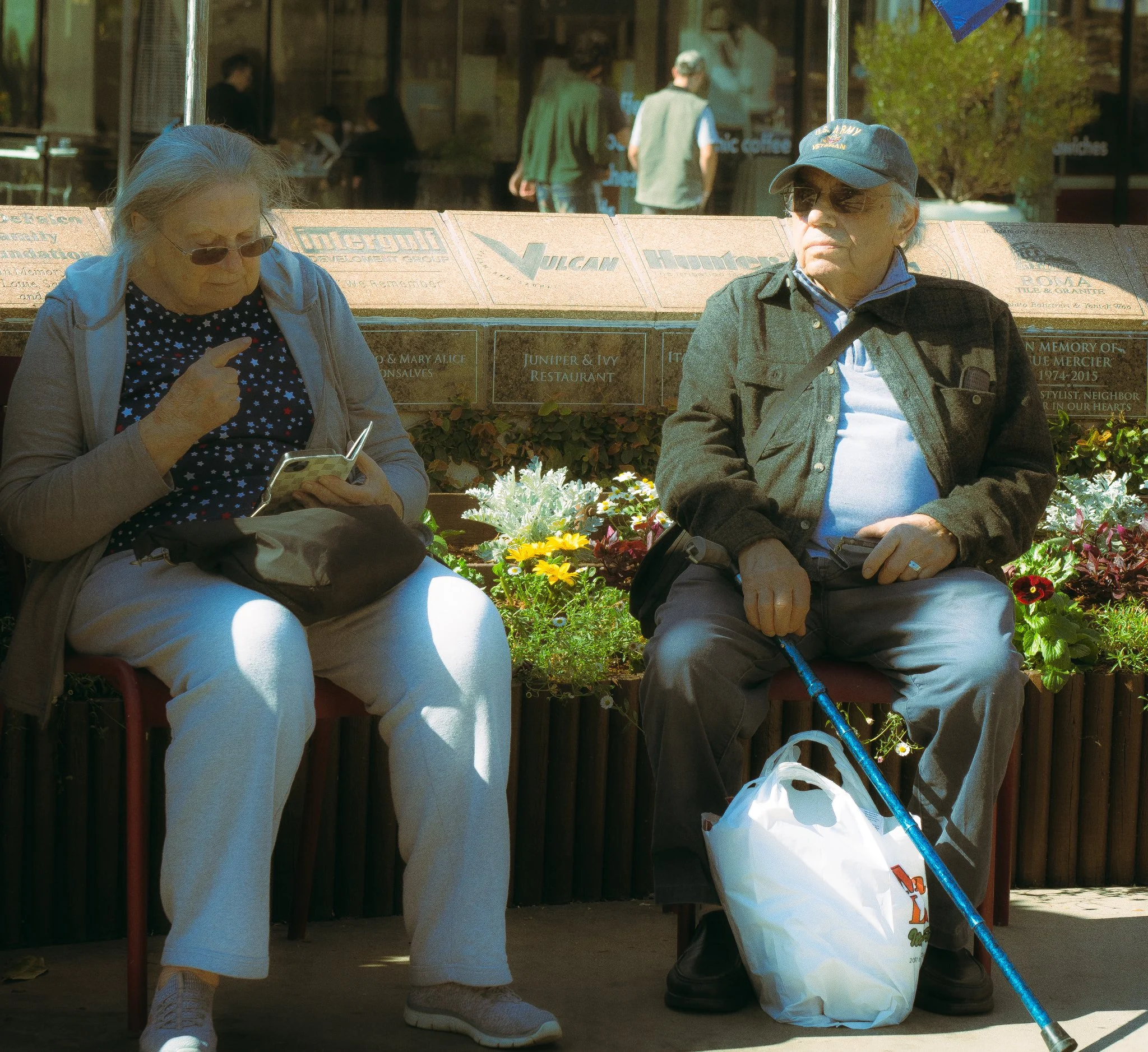 Two elderly people sitting on a bench outdoors, one woman looking at her phone and one man with sunglasses and a cap, with trash bags and a walking stick. Behind them is a glass storefront and a flower bed.
