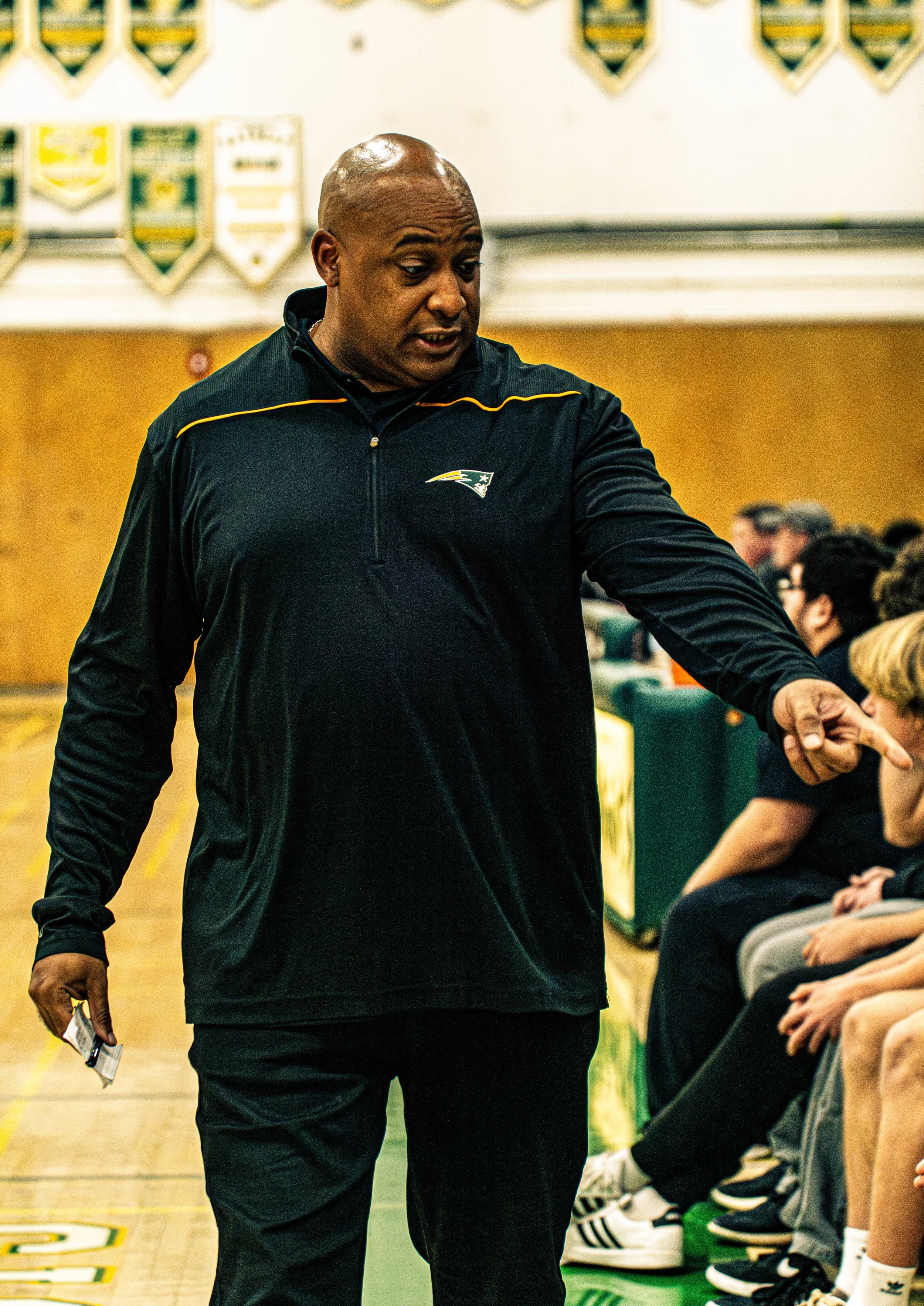 A man dressed in a New England Patriots jacket is standing and talking to a group of young people sitting on chairs in a gymnasium. The background features banners and a white wall.