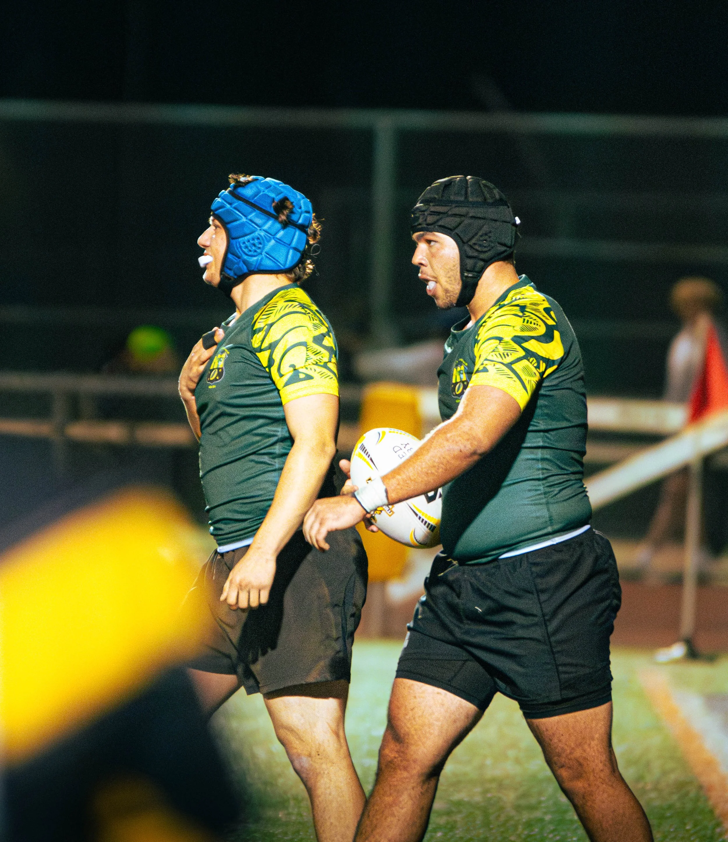 Two rugby players in black and yellow uniforms, wearing protective headgear, stand on a field during a match, one holding a rugby ball.
