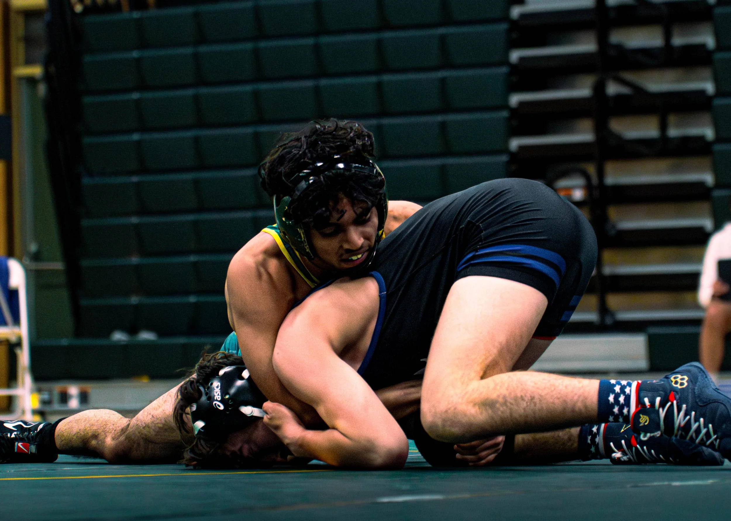 Two wrestlers engage in a match on the mat in an indoor arena, with one wrestler on top and holding the other in a hold.