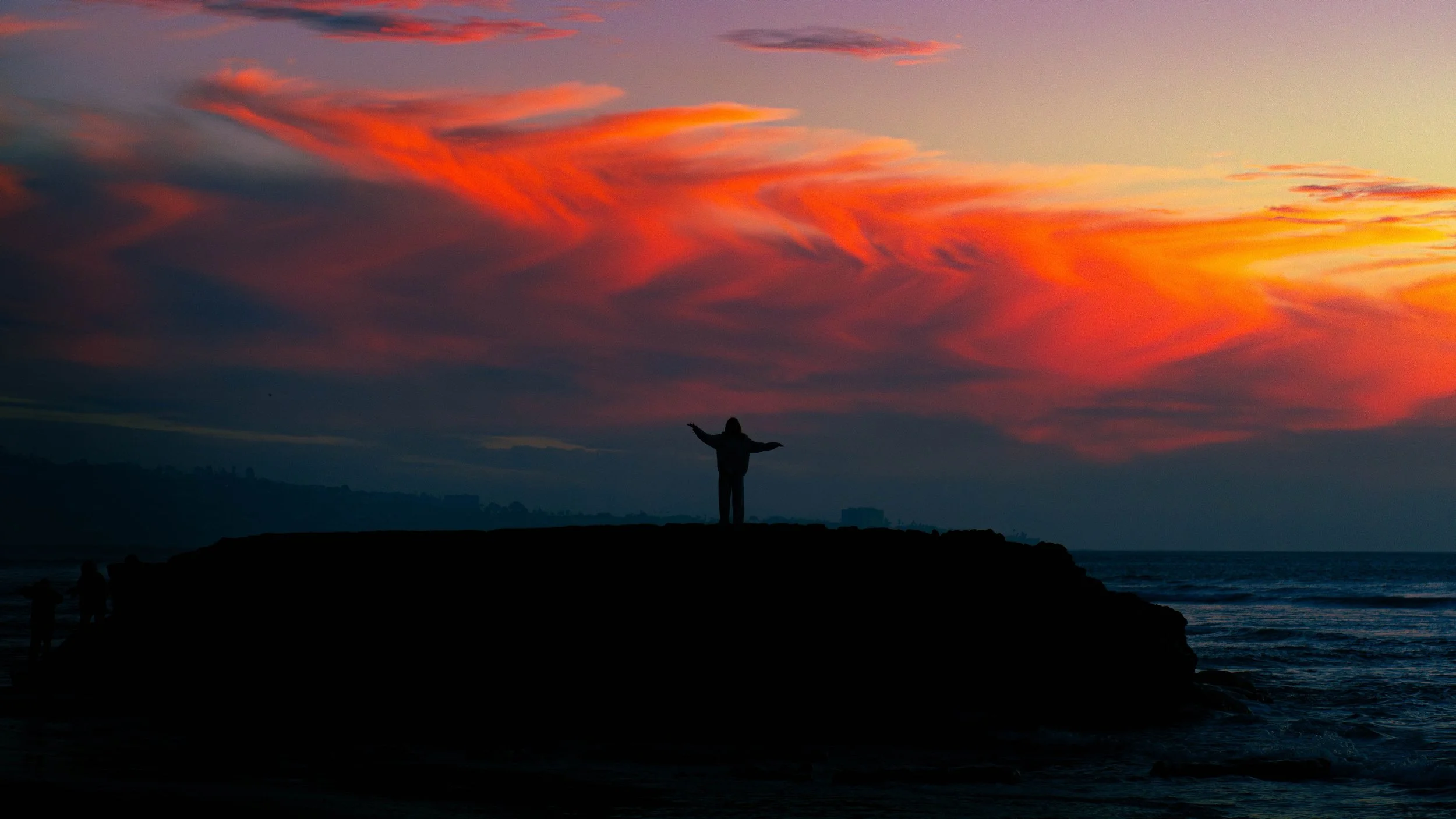 Person standing on a rocky outcrop with arms outstretched, silhouetted against a colorful sunset sky over the ocean.