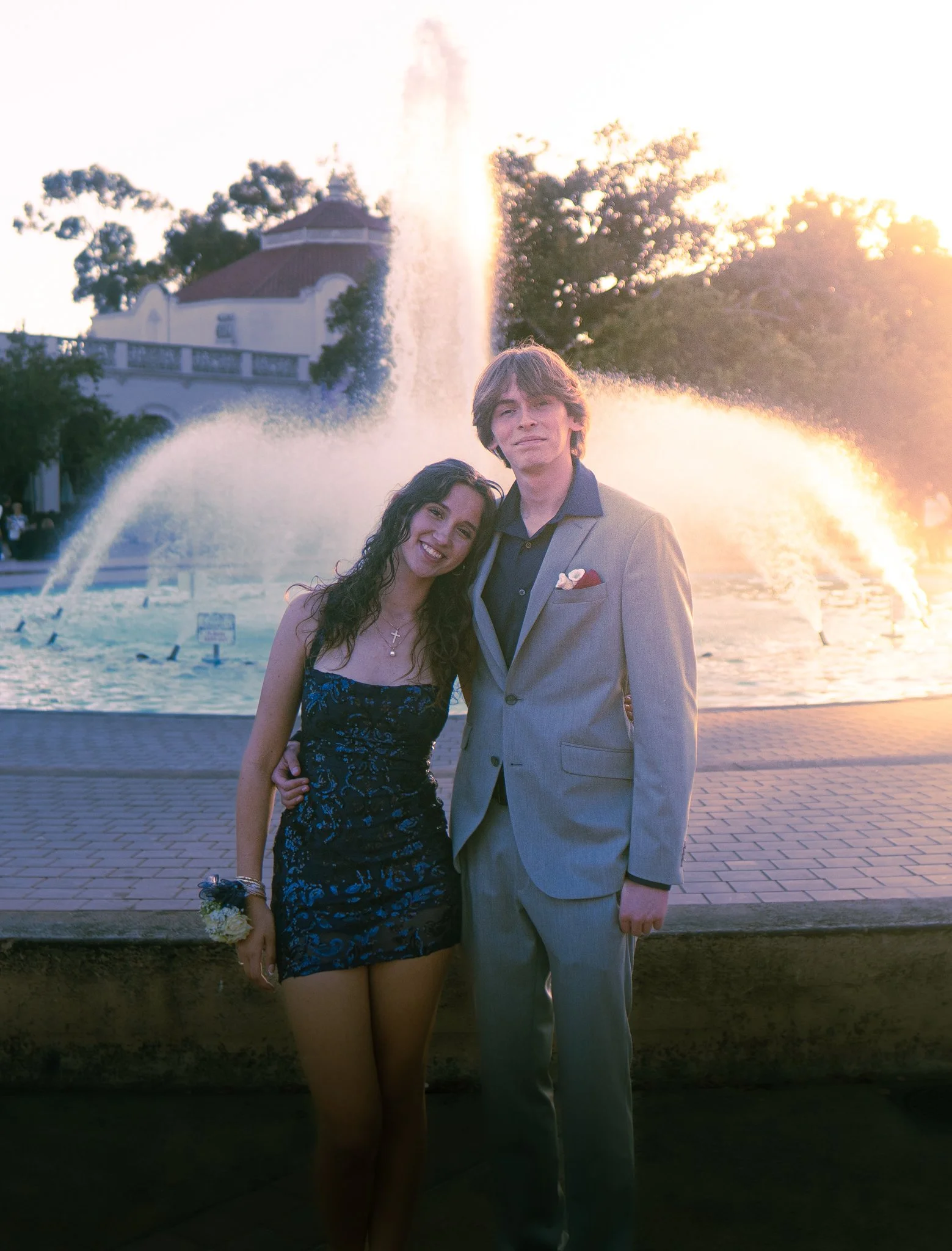 A young woman and a young man standing close together outdoors during sunset, smiling in front of a fountain and a building with a dome, surrounded by trees.