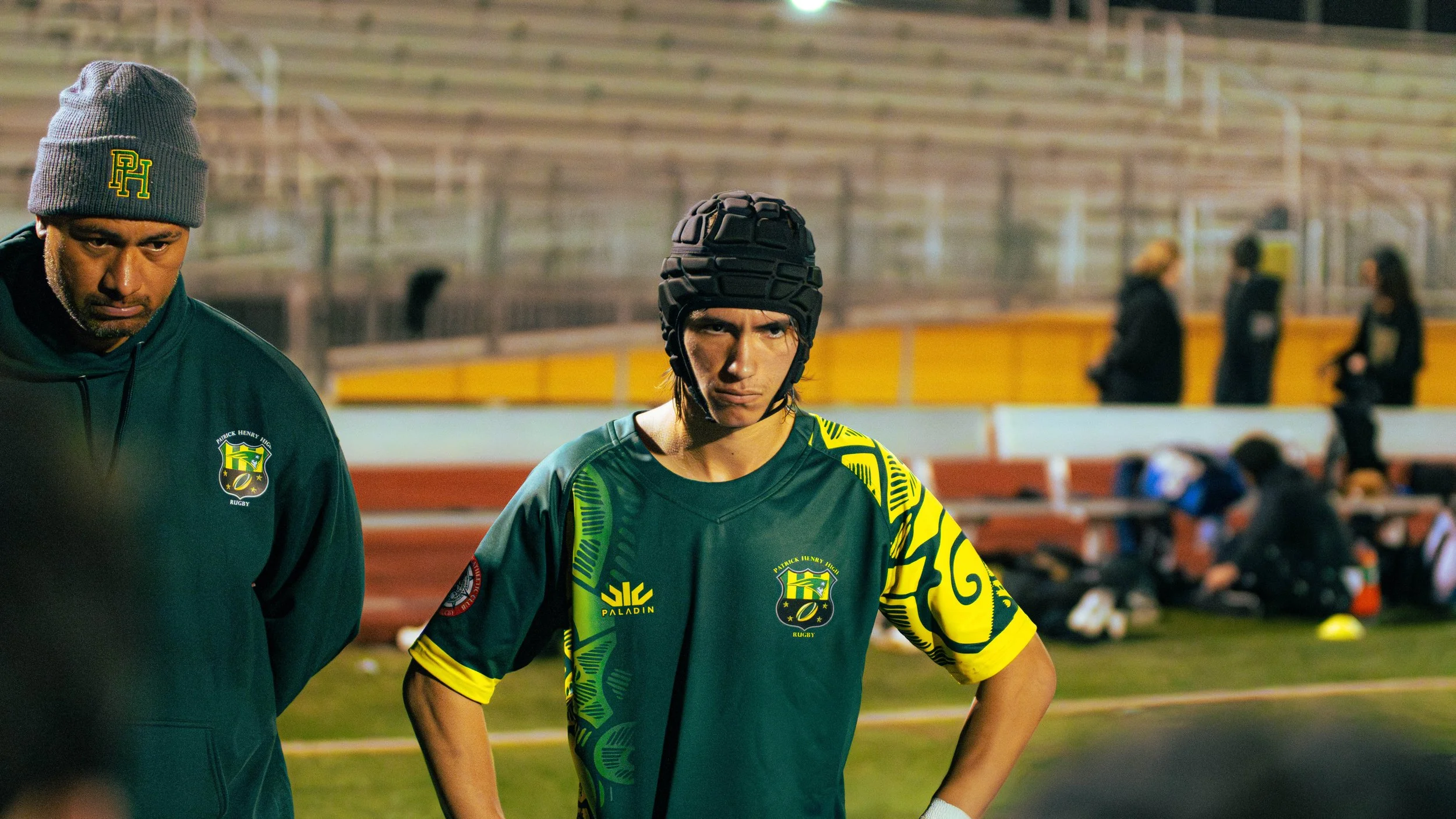 Two rugby players dressed in green and yellow uniforms standing on a field in a stadium, with a blurred background of a few people sitting on benches.