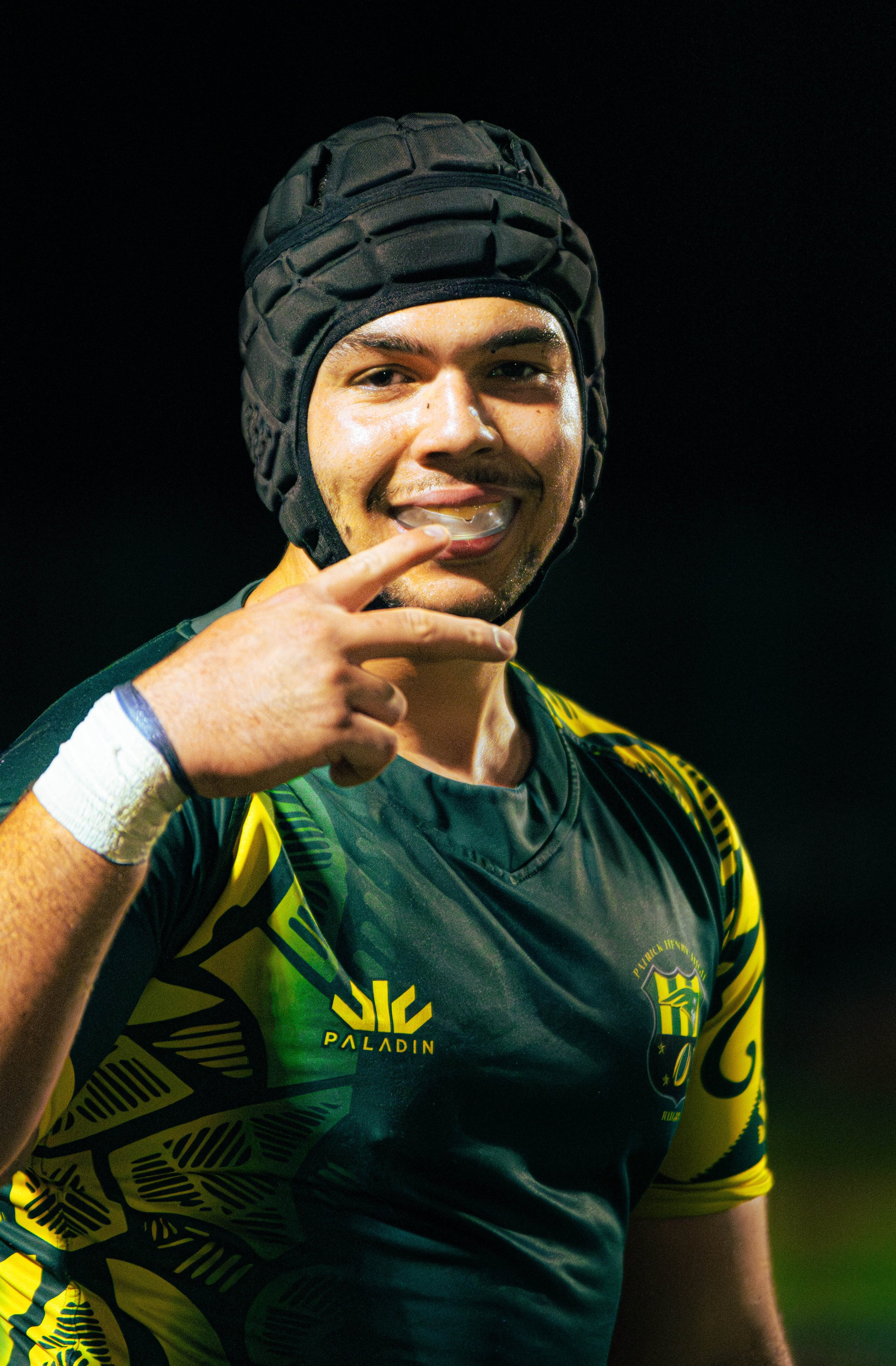 Young male rugby player wearing yellow and black uniform, black padded headgear, smiling, touching his chin, dark background.