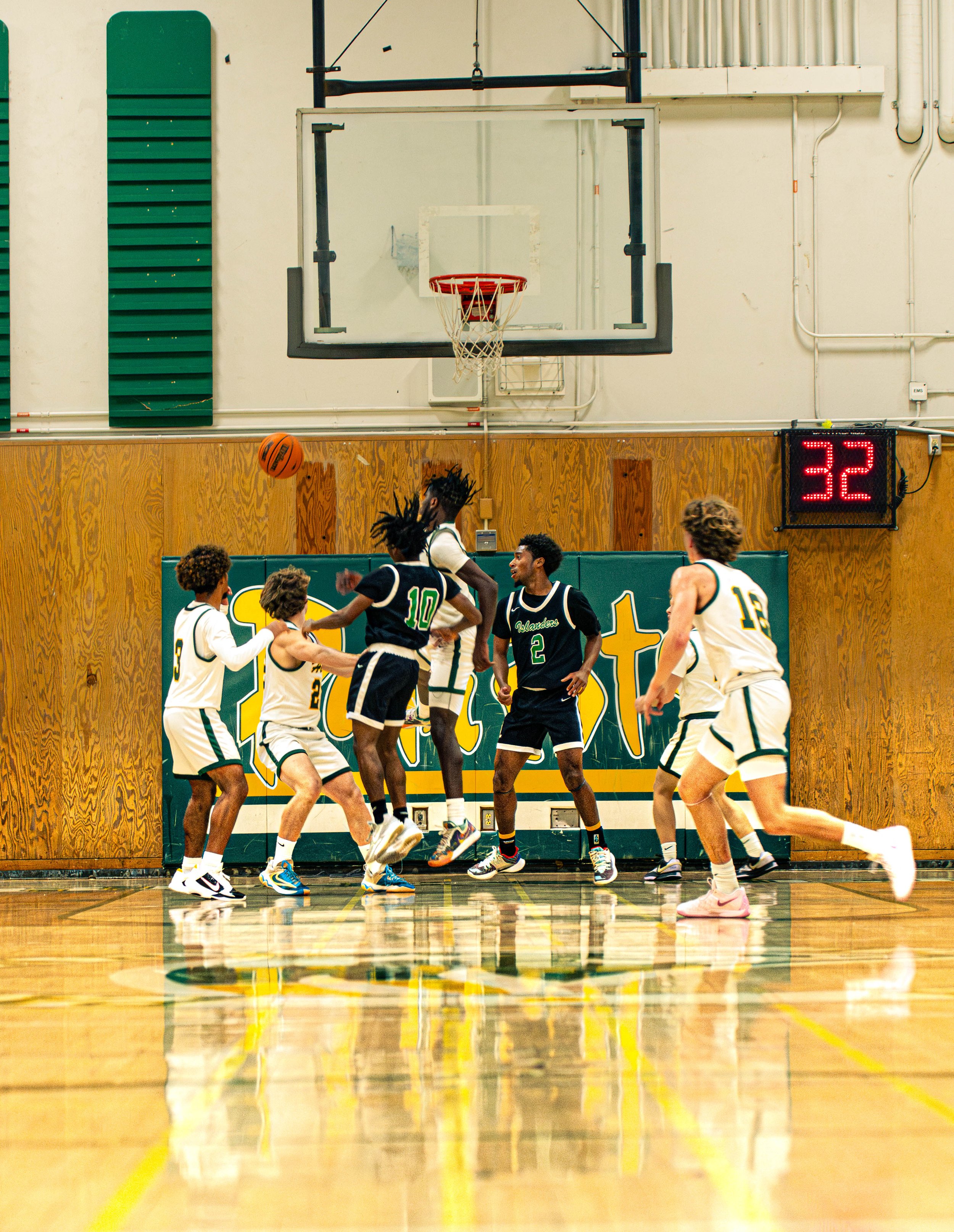 A group of basketball players in white and black uniforms play on a wooden court inside a gymnasium with a scoreboard displaying the number 32.