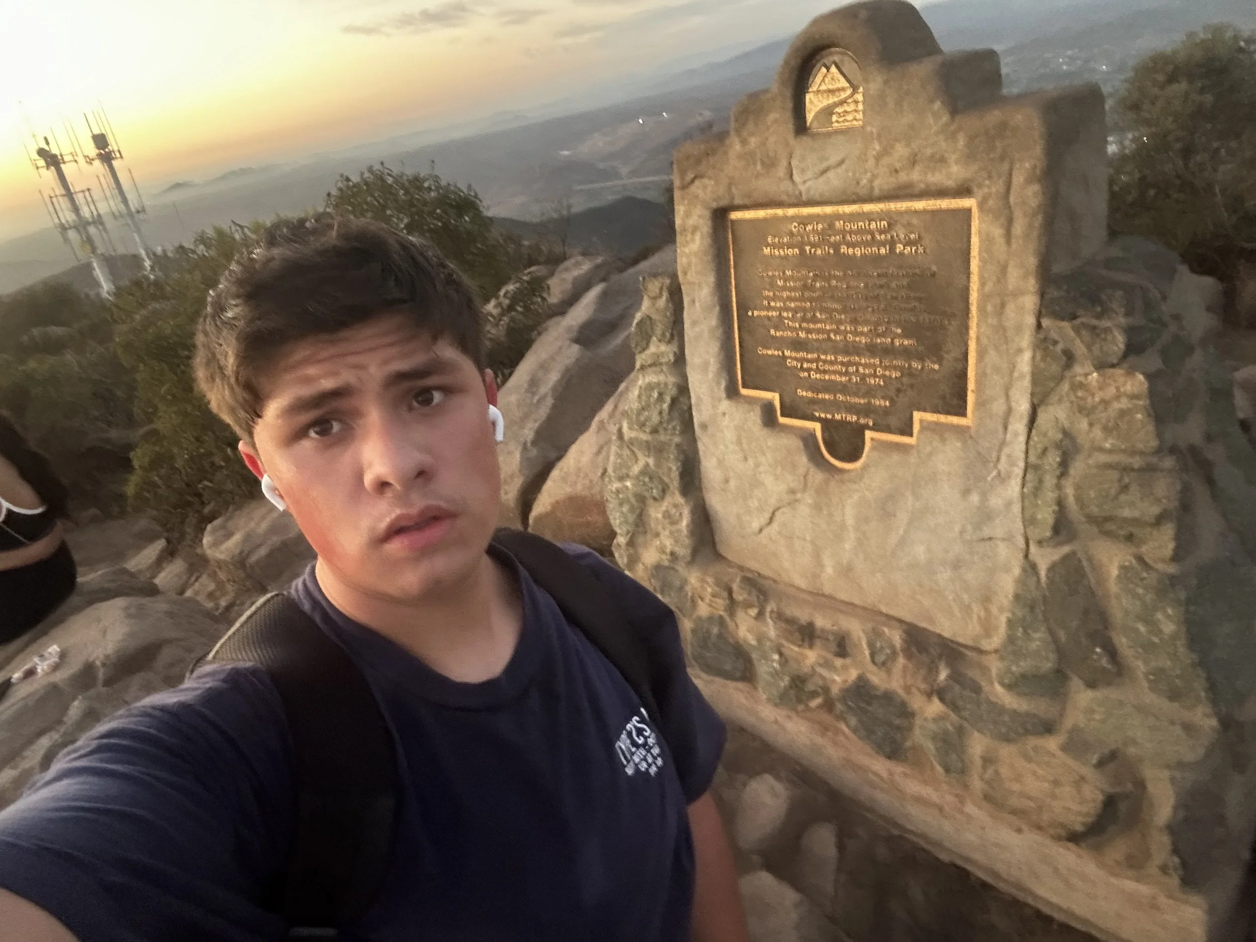 A young man with dark hair, wearing a dark t-shirt and wireless earbuds, taking a selfie at sunset on a mountain trail with a plaque and communication towers in the background.
