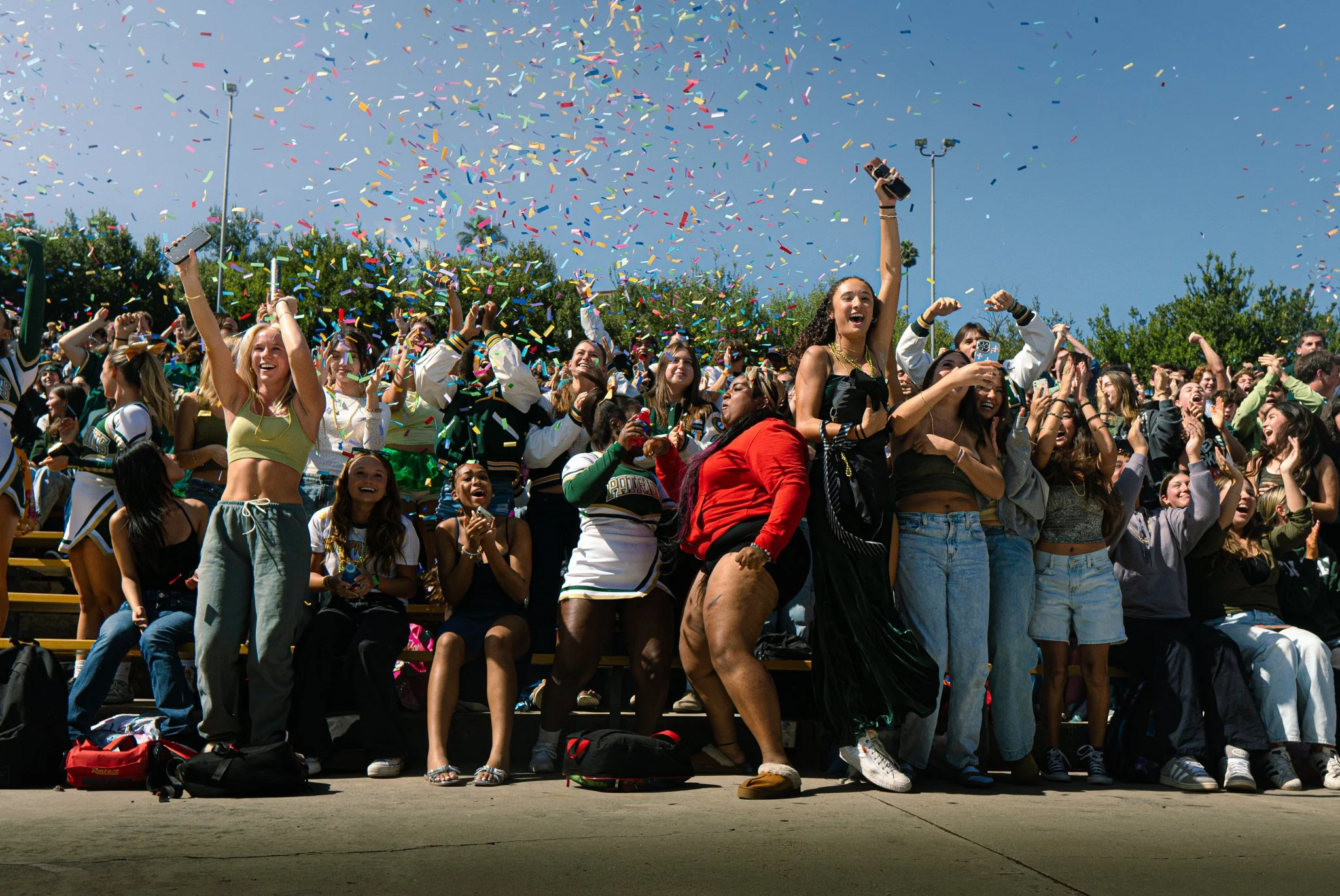 A group of people gathered outdoors under a clear blue sky, celebrating with confetti falling. Some are standing and cheering, holding phones, and wearing sports apparel, indicating a festive or sports-related event.