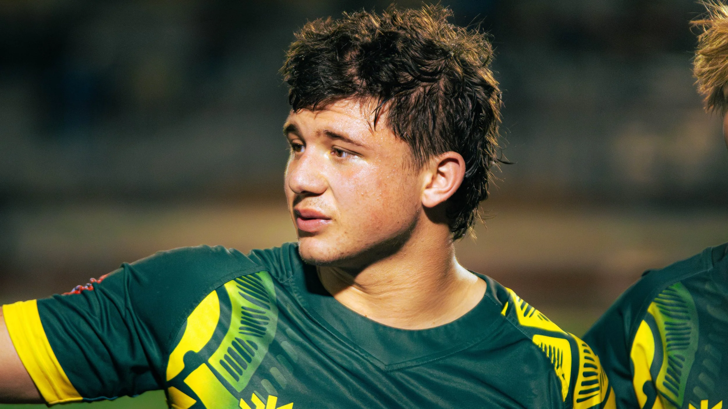 Close-up of a young male soccer player with curly hair wearing a green and yellow jersey, looking to the side after a game.