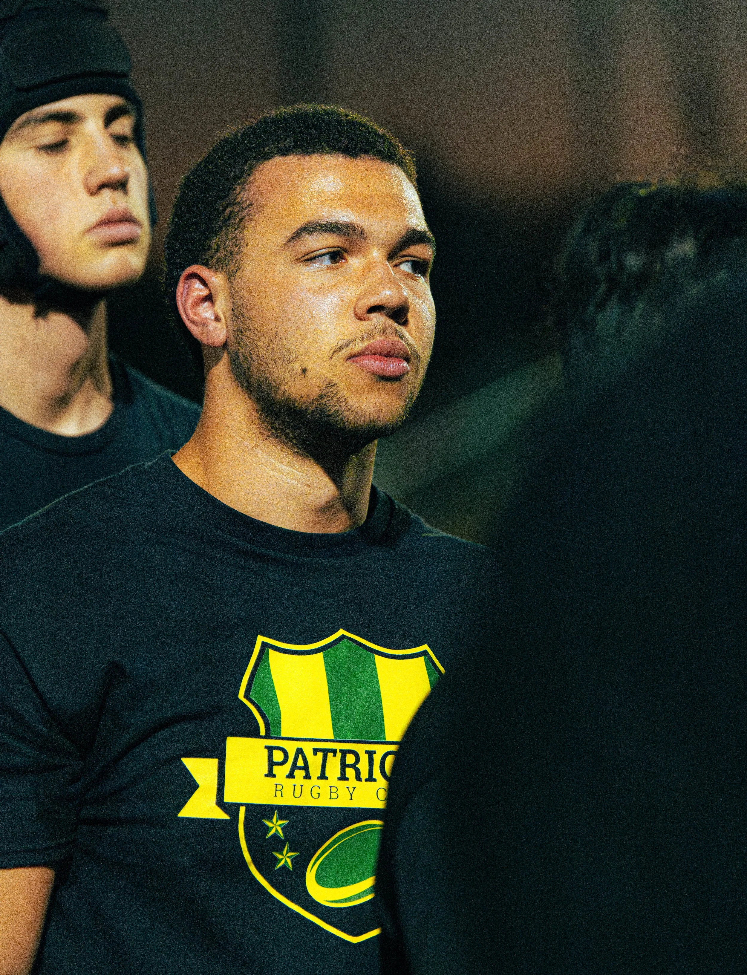 A young man wearing a black shirt with a yellow and green logo of Patrico Rugby Club, standing next to someone in a rugby helmet, with a third person partially visible on the right.