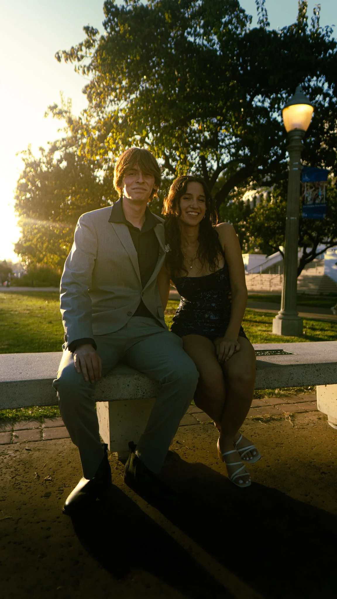 A man and woman sitting on a concrete bench outdoors during sunset, with trees and a lamp post in the background.