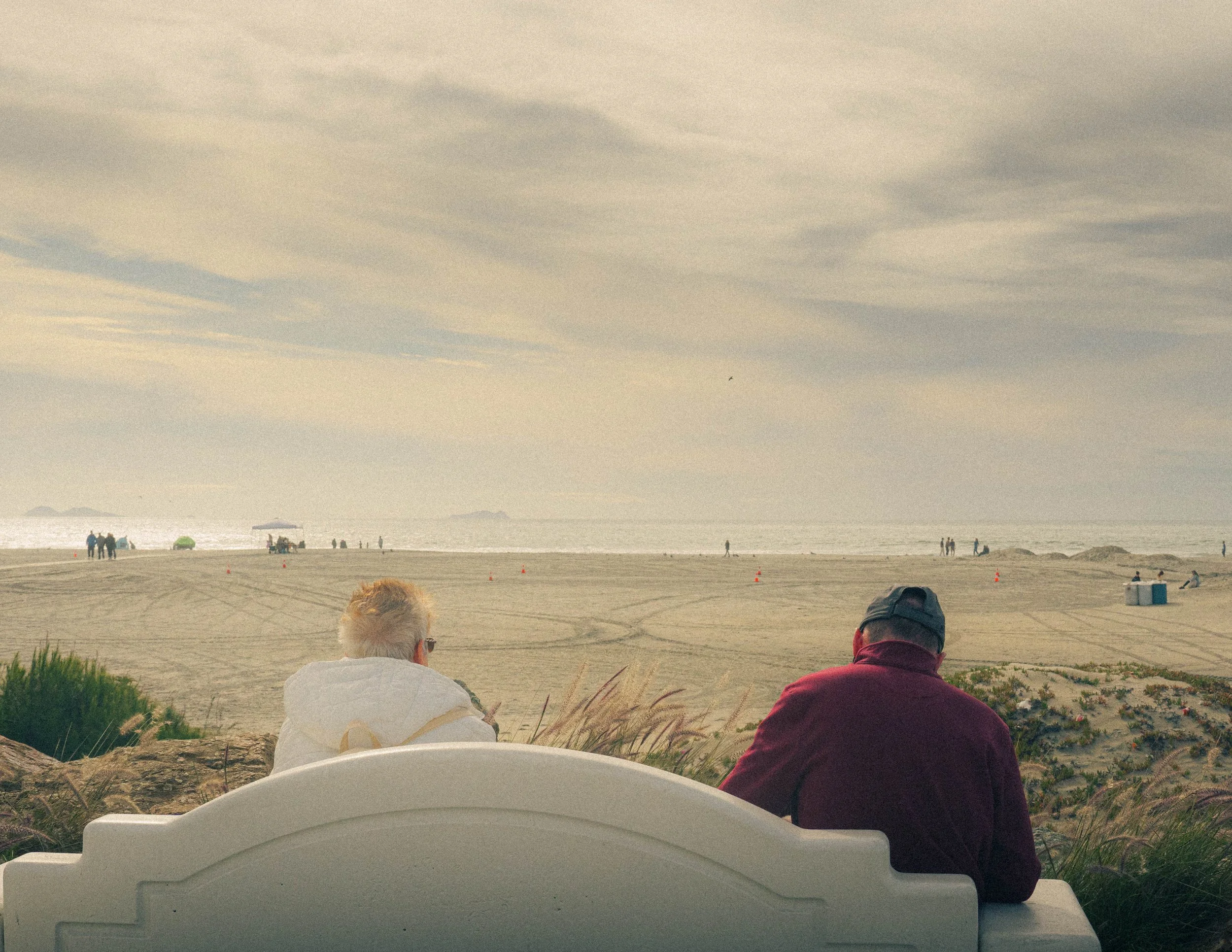People sitting on the sandy beach facing the ocean with cloudy sky overhead and a few distant boats or ships in the water.