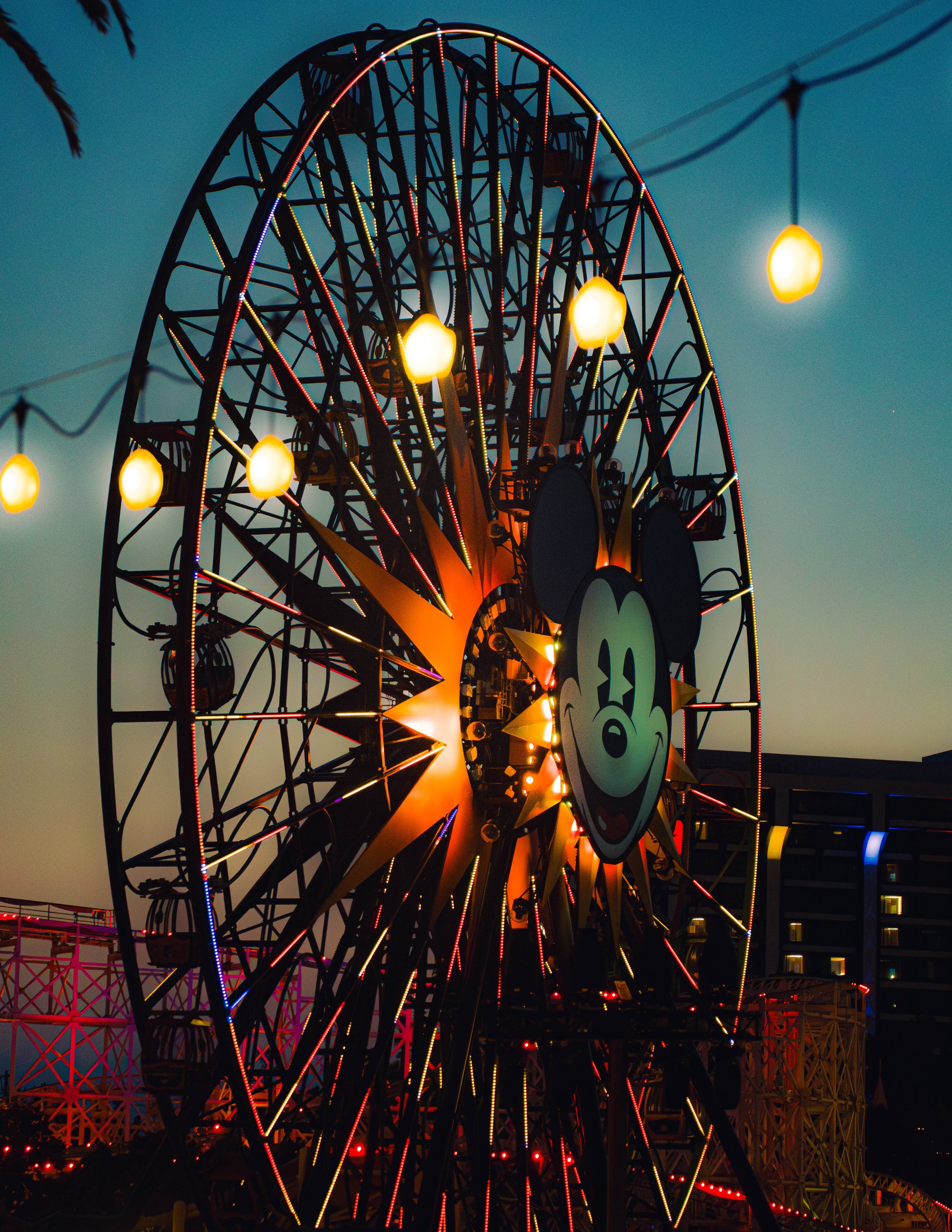 A brightly lit Ferris wheel with Mickey Mouse's face on it, set against the evening sky at a carnival.