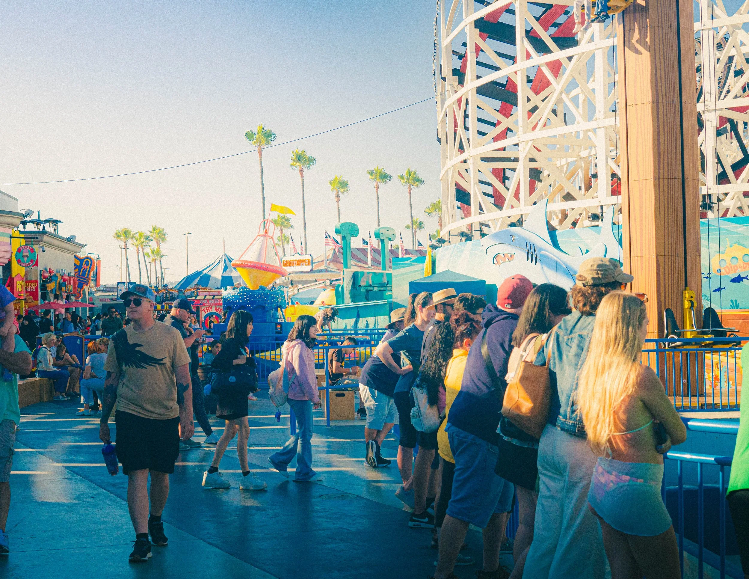 A crowded amusement park or fair with people walking and queuing, a roller coaster track, and palm trees under a clear sky.
