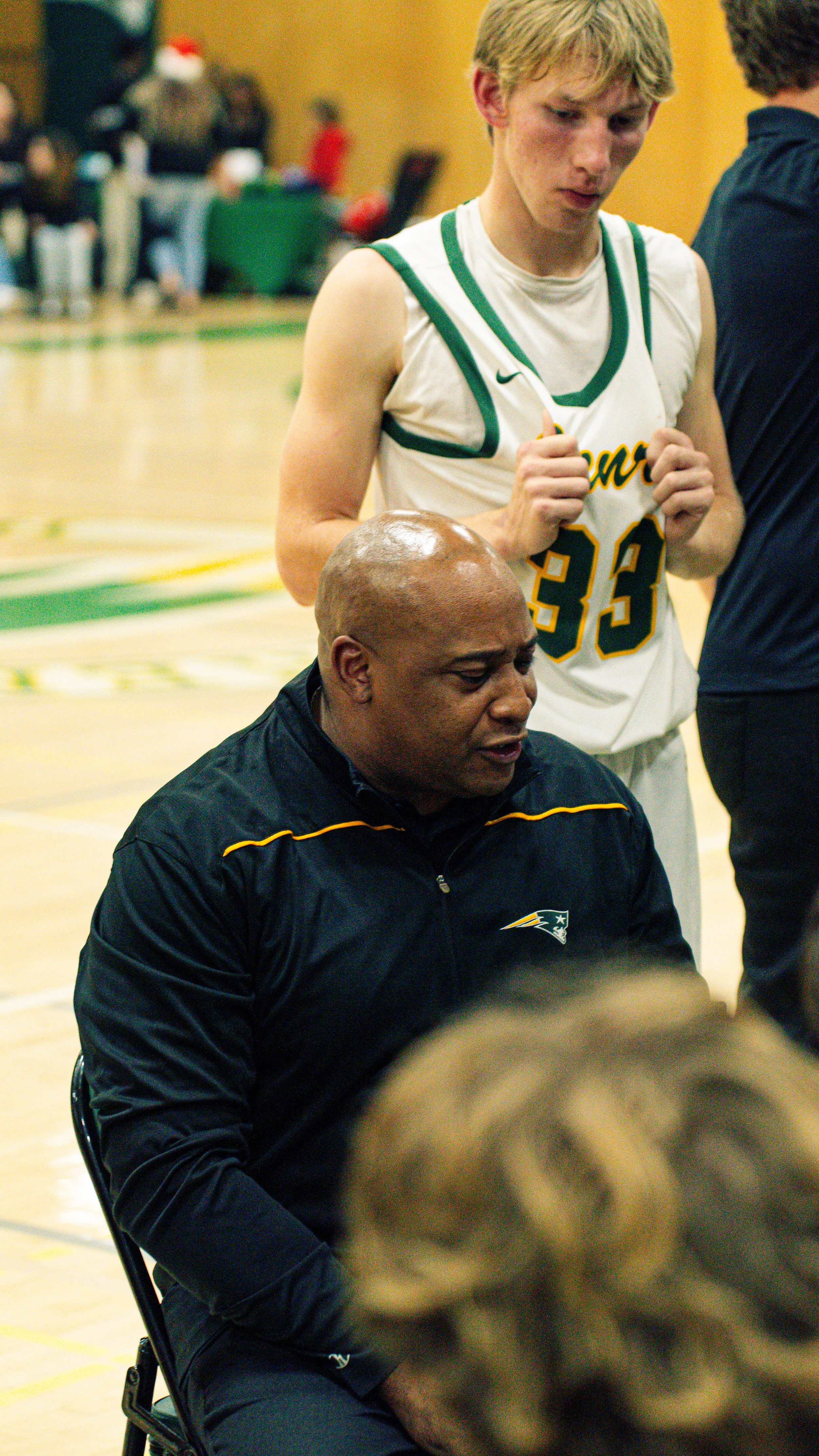 A young male basketball player with blonde hair and wearing a white and green uniform is standing next to a seated man in a black tracksuit with a Patriots logo. They are on a basketball court, with a blurred crowd of spectators in the background.