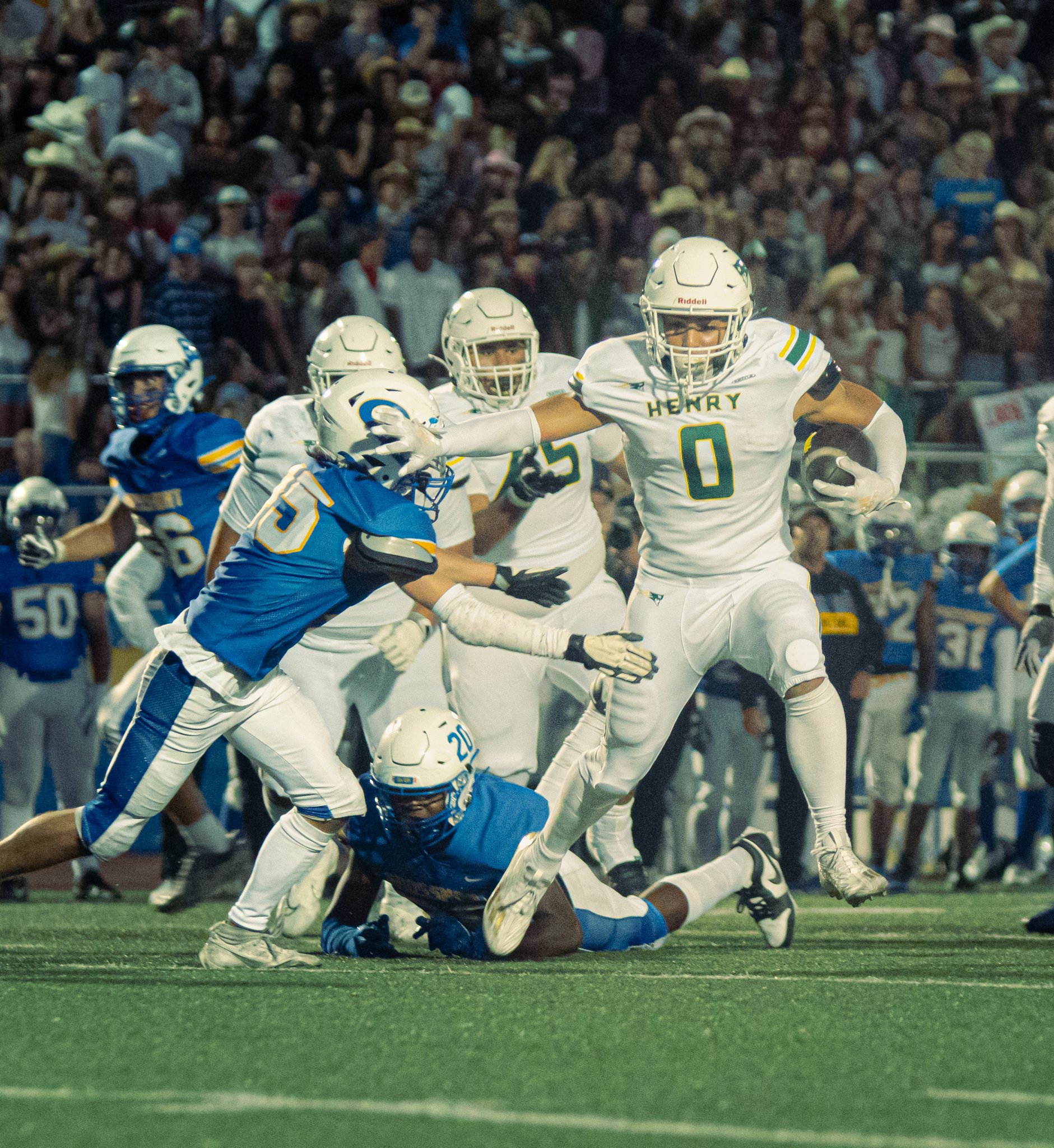 Football players in white and blue jerseys are in action during a game, with one player in a white jersey carrying the ball and others trying to tackle him. Crowd of spectators in the background.