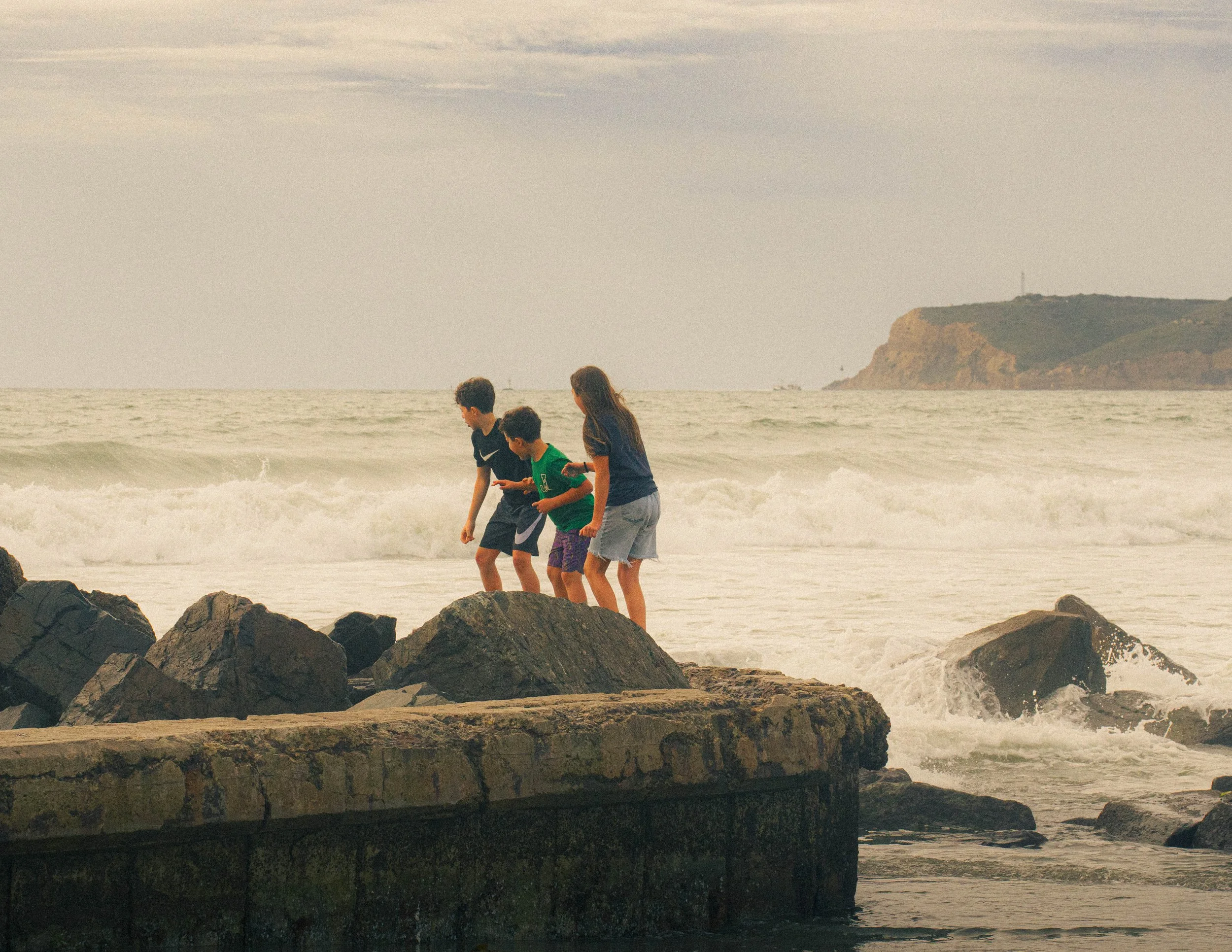 Three children playing near rocks on a beach, with ocean waves and a distant cliff in the background.
