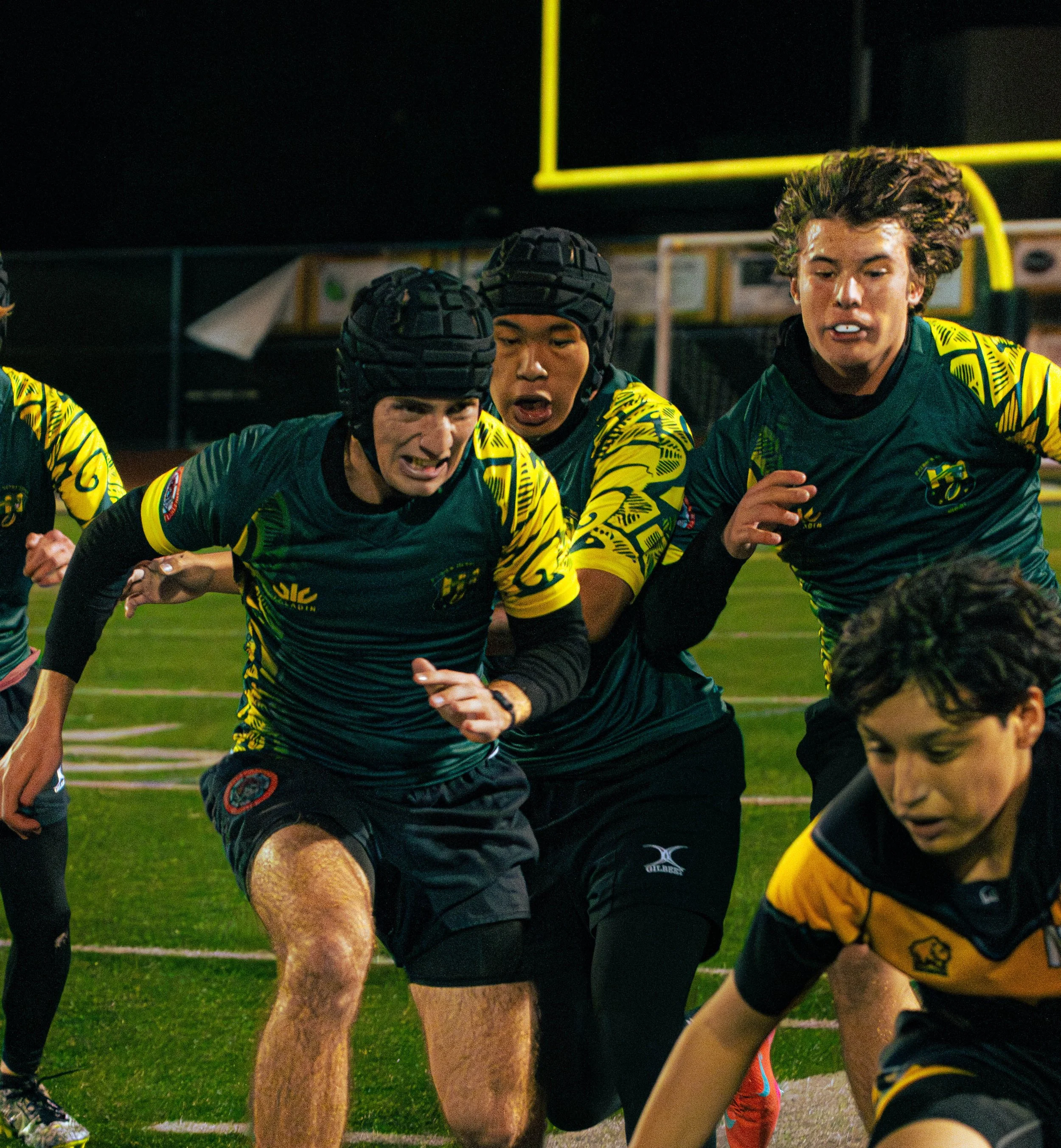Group of young male rugby players in yellow and green uniforms, wearing helmets, crouched down during a game on a football field at night.