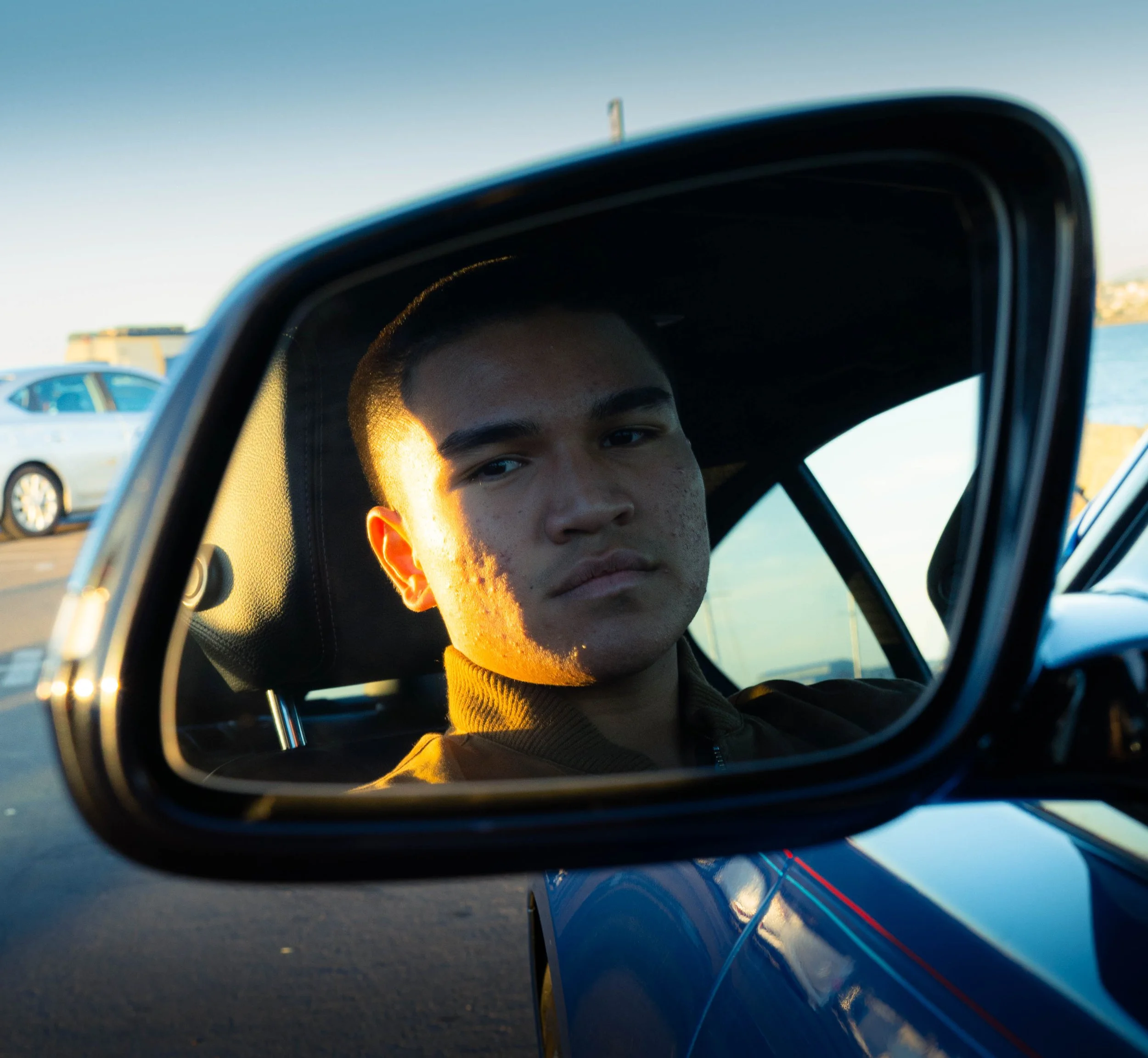 Close-up of a young man's face reflected in a car's side mirror during sunset, with part of the car visible in the frame and a parking lot with other cars in the background.