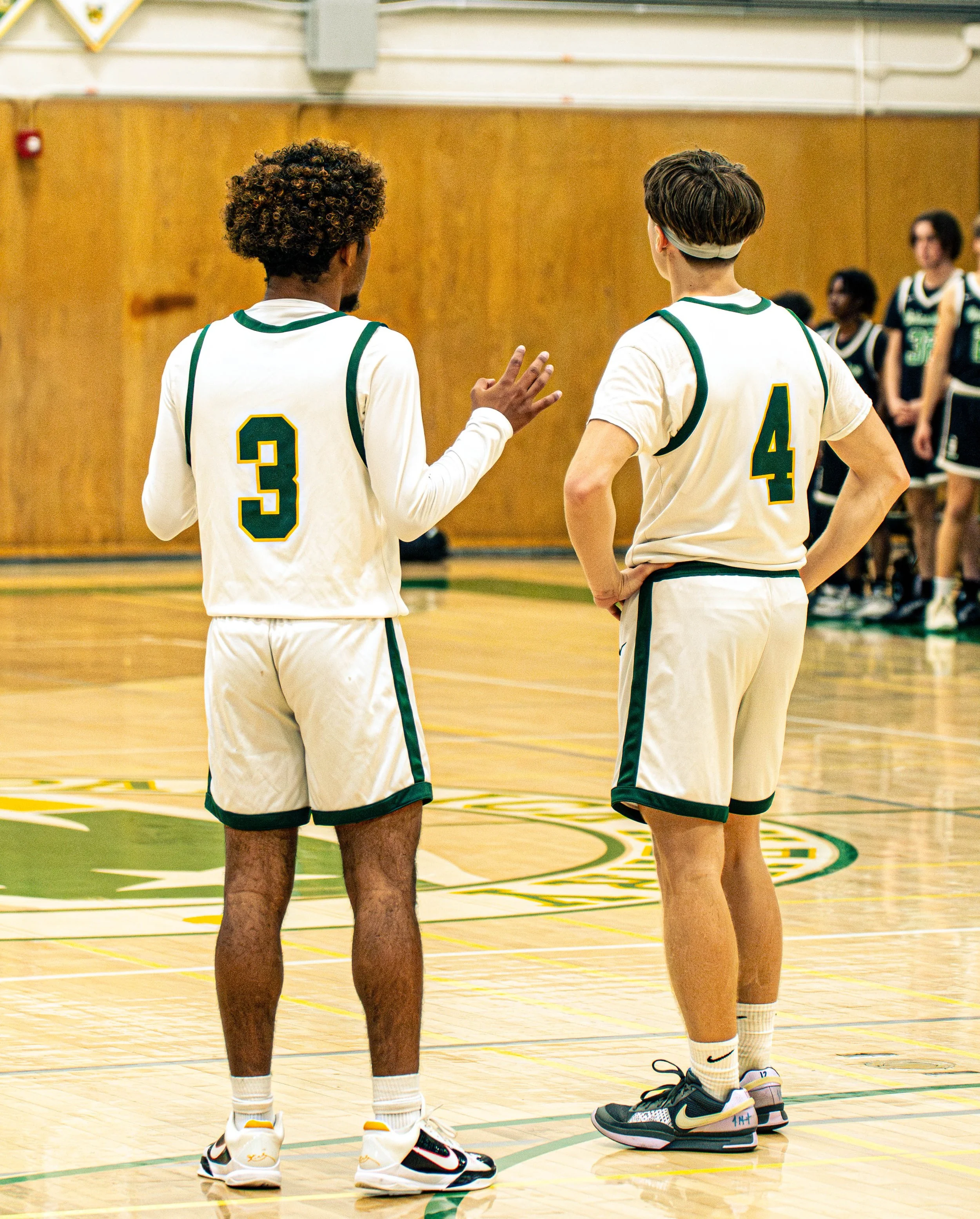 Two basketball players, wearing white jerseys with green and yellow trim and shorts, stand on a gymnasium court with one player gesturing with his hand as they talk. Several other players are visible in the background, standing against a wooden wall.