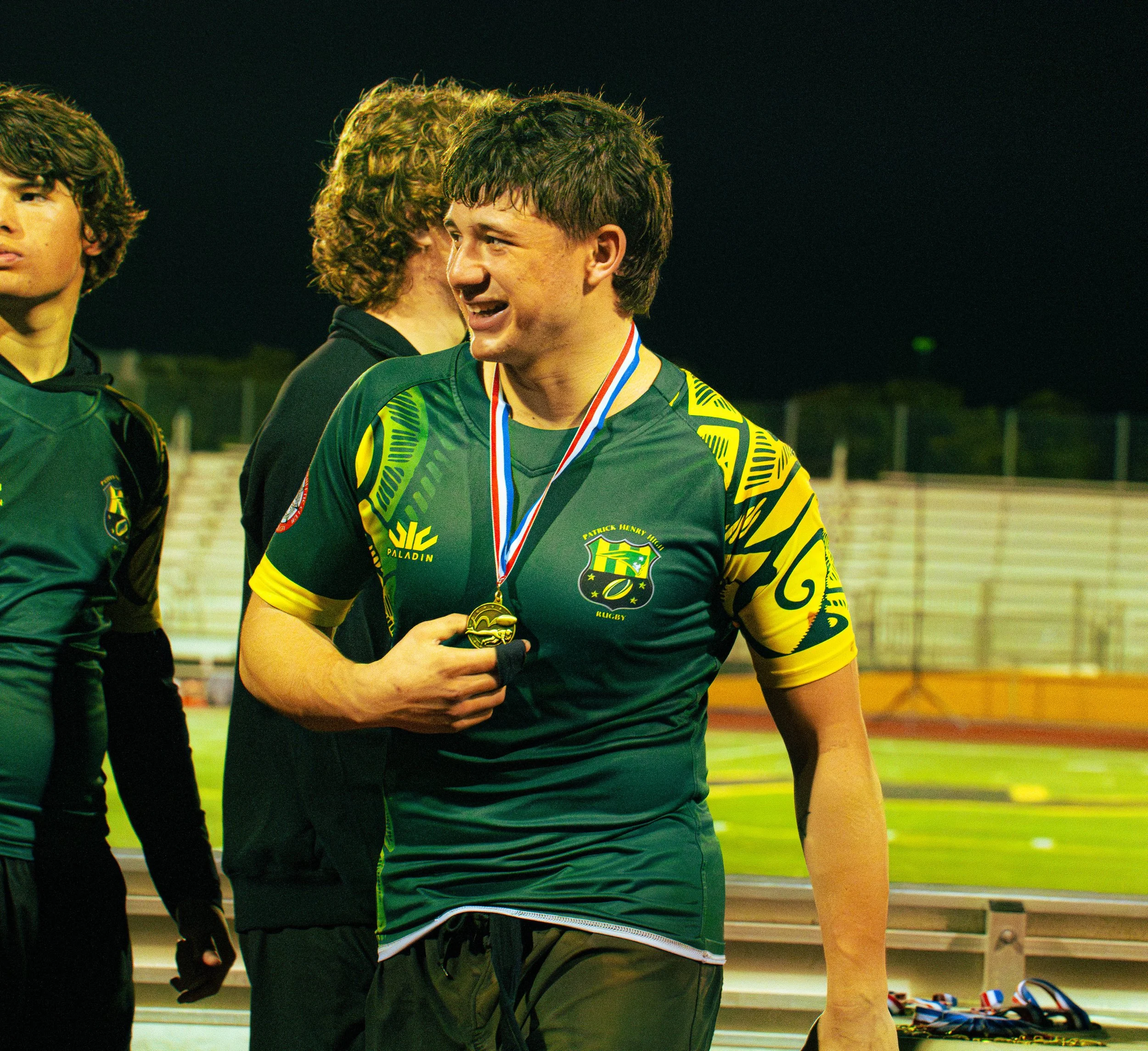 Three young men in dark athletic uniforms standing on a sports field at night, with medals around their necks, celebrating a victory.