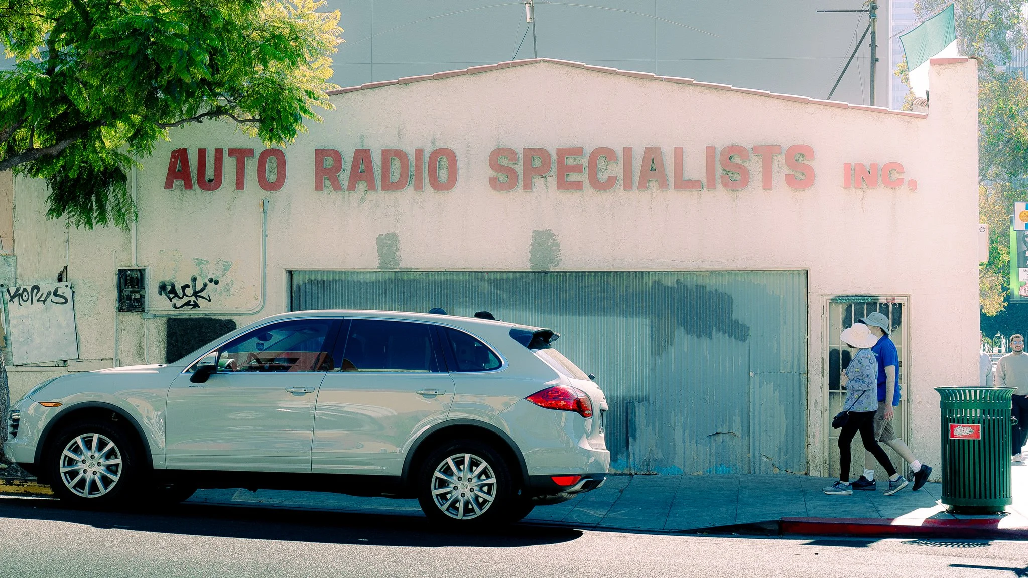 Street scene with a silver car parked in front of a building with a sign that reads 'AUTO RADIO SPECIALISTS INC.' Two pedestrians wearing hats walk past a green trash bin on the sidewalk. A green tree partially obscures the building, and graffiti is 