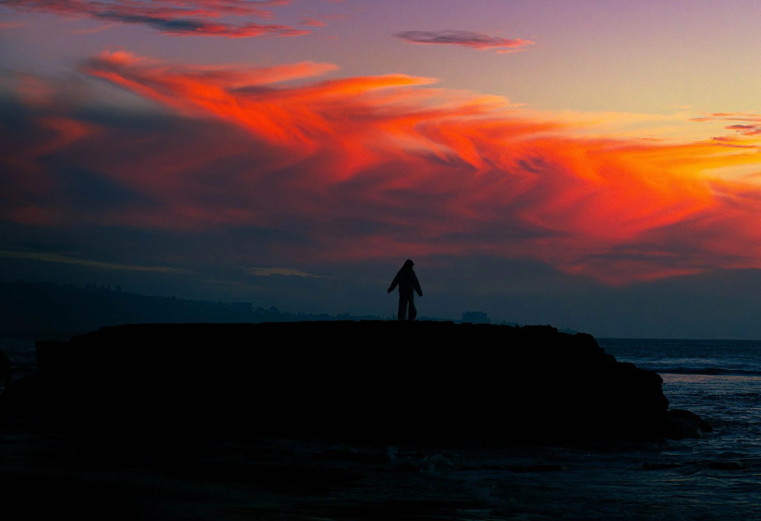 A silhouetted person standing on a rocky cliff overlooking the ocean at sunset, with colorful pink and orange clouds in the sky.