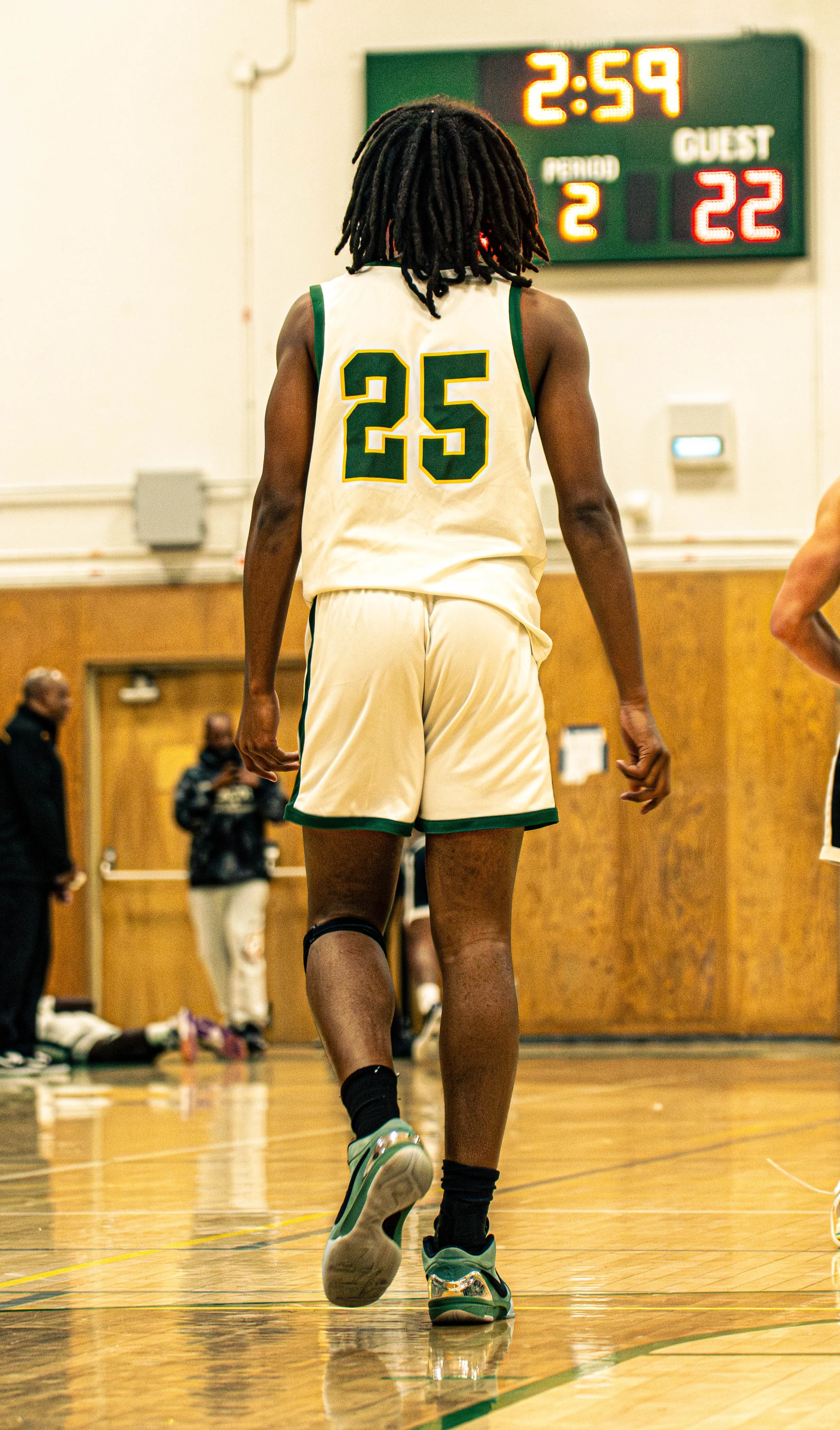 A basketball player wearing a white jersey with green and yellow accents, number 25, walking on an indoor basketball court. The digital score display shows 2:59 and a score of 22-2. People are visible in the background.