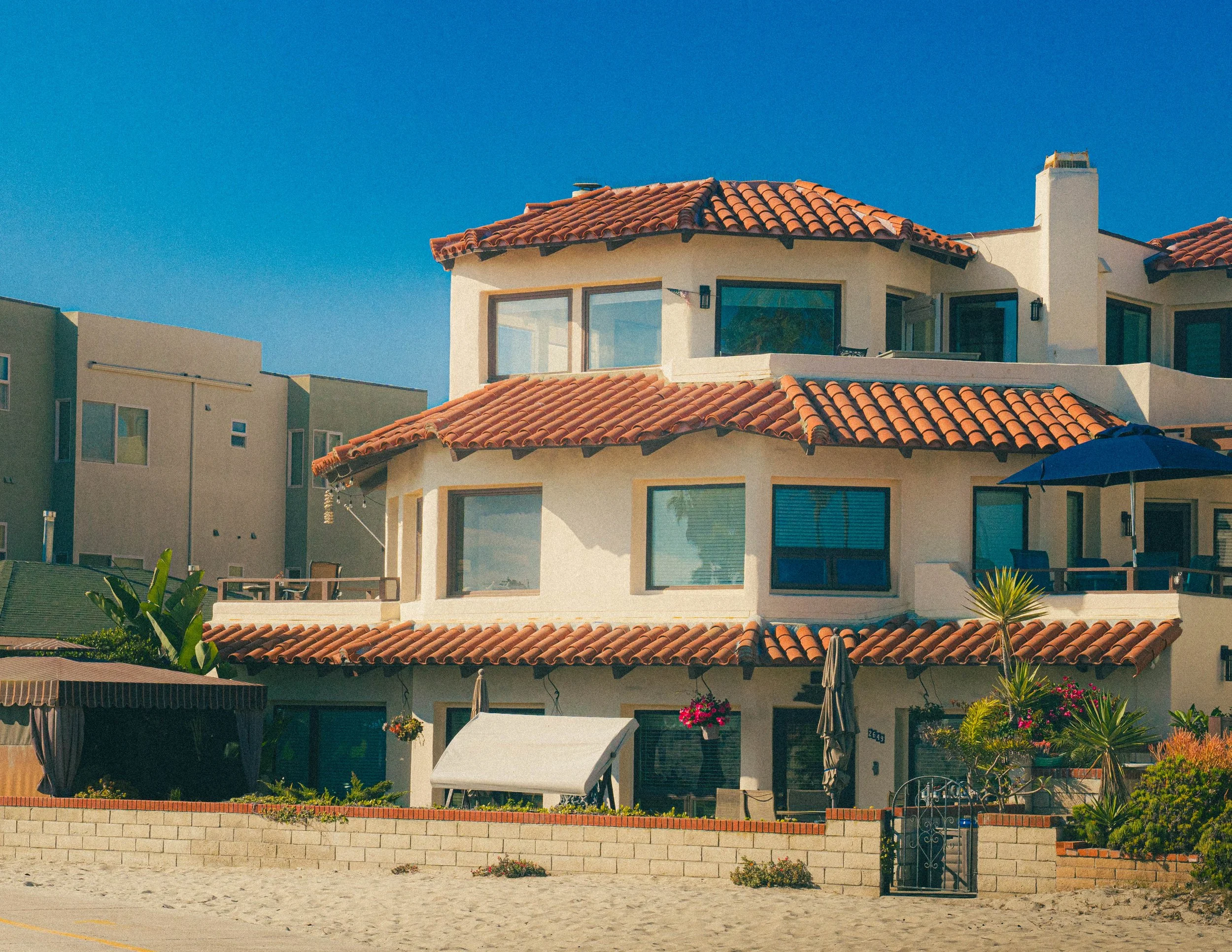 A multi-story house with a red-tiled roof, multiple large windows, a chimney, and outdoor patio furniture, set against a clear blue sky.